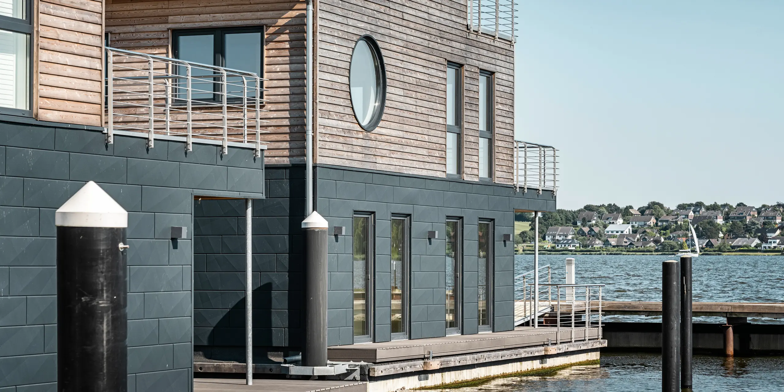 Nahaufnahme eines stilvollen Wasserhauses im Pionierhafen Schleswig mit einer Fassade aus PREFA Siding.X in P.10 Anthrazit und natürlichem Holz am oberen Stockwerk. Ein rundes und mehrere eckige Fenster sowie klare Linien an der Konstruktion bieten einen modernen Kontrast zur traditionellen Landschaft im Hintergrund. Diese schwimmenden Häuser sind mit ihren nachhaltigen Materialien und direktem Zugang zum Wasser ausgestattet. Die elf Wasserhäuser in Schleswig sind ein Beispiel für modernes Wohnen in Harmonie mit einer maritimen Umgebung.