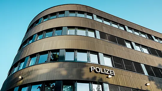 Dramatische Untersicht des geschwungenen Neubaus vom Polizeikommissariat 43 in Hamburg: Die markante PREFA Aluminiumfassade aus eloxierten Strangpressprofilen „Zacke“ in Mittelbronze zieht als architektonischer Eyecatcher alle Blicke auf sich. Die feine, rhythmische Struktur erzeugt dynamische Licht- und Schattenverläufe und betont die fließenden Rundungen des Baukörpers. PREFA Aluminium überzeugt durch hohe Witterungsbeständigkeit, Korrosionsschutz und Langlebigkeit – eine nachhaltige, wartungsarme Lösung für präzise, moderne Fassadenarchitektur.