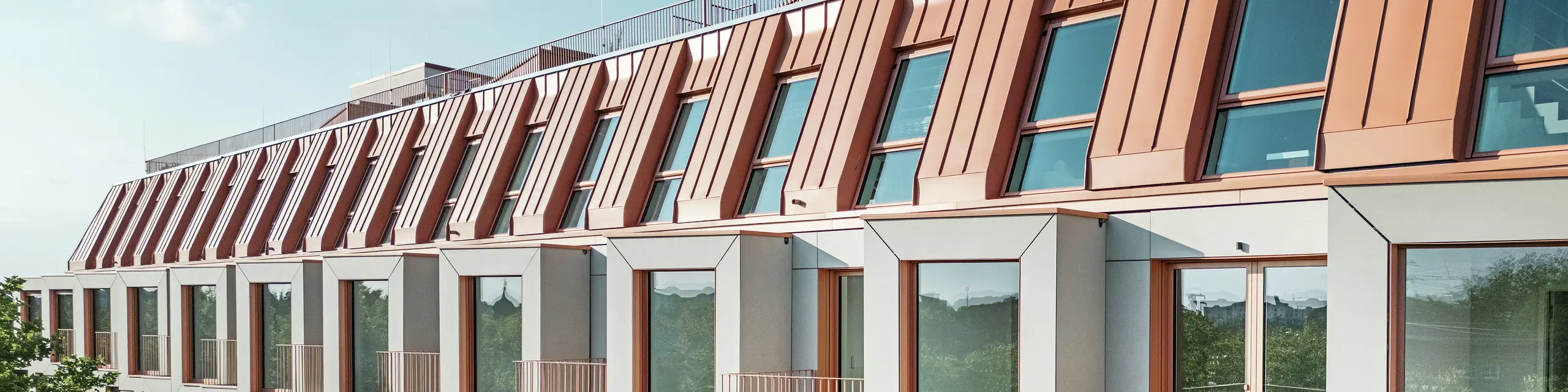 Facade view of the Van B residential project in Munich with the striking PREFALZ aluminium roof in the special colour IGP Orange HDP from PREFA. The modern roof covering combines aesthetic elegance with technical precision and offers lasting protection against the elements. The cubic bay windows with generous window areas and filigree balconies create a harmonious interplay of light, space and functionality – ideal for sophisticated living concepts in urban environments.