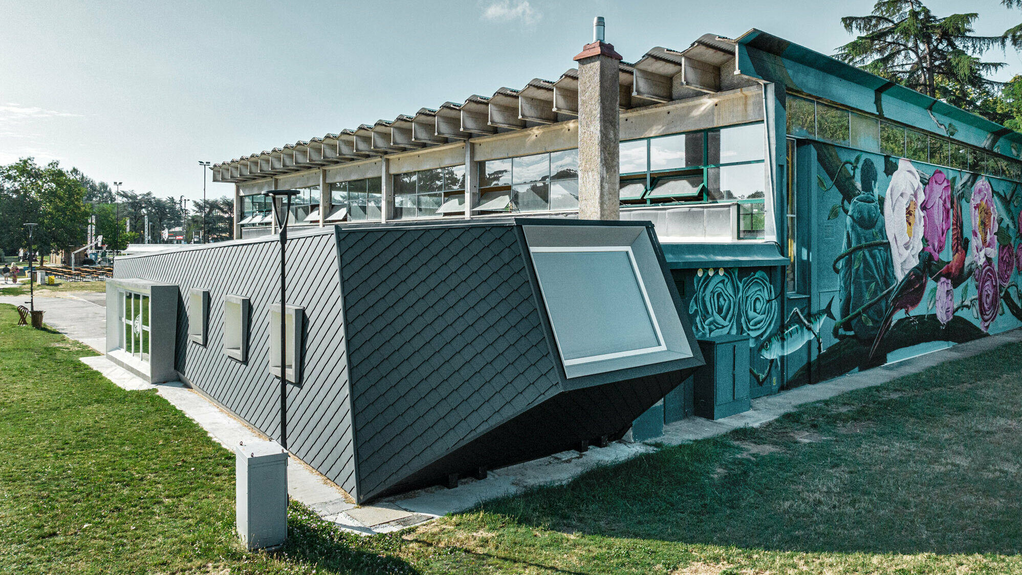 The Rodriguez gym and its new extension with the scaly shomboid skin made of PREFA aluminium from the side, the lawn extending before it and on the side. Part of a sports field can be seen in the background.