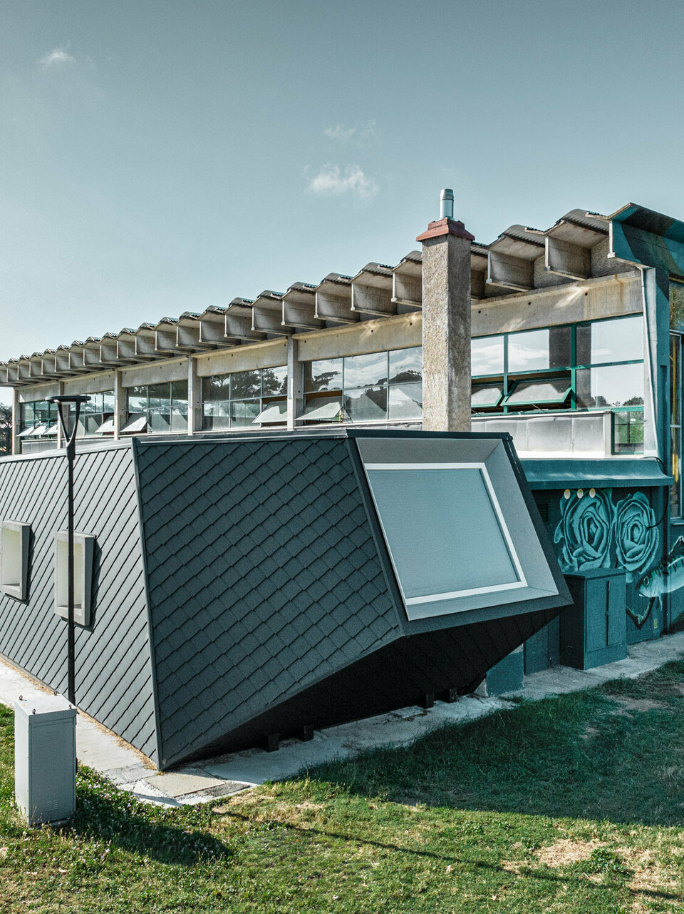 The Rodriguez gym and its new extension with the scaly shomboid skin made of PREFA aluminium from the side, the lawn extending before it and on the side. Part of a sports field can be seen in the background.