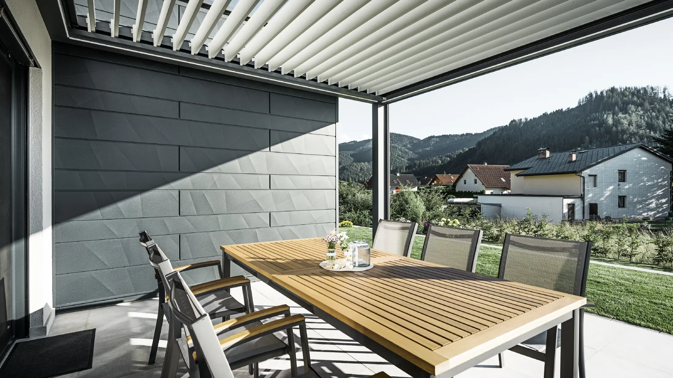 Sheltered terrace covered with slats, with table and chairs; one side of the façade is clad with PREFA Siding.X façade panels in anthracite.