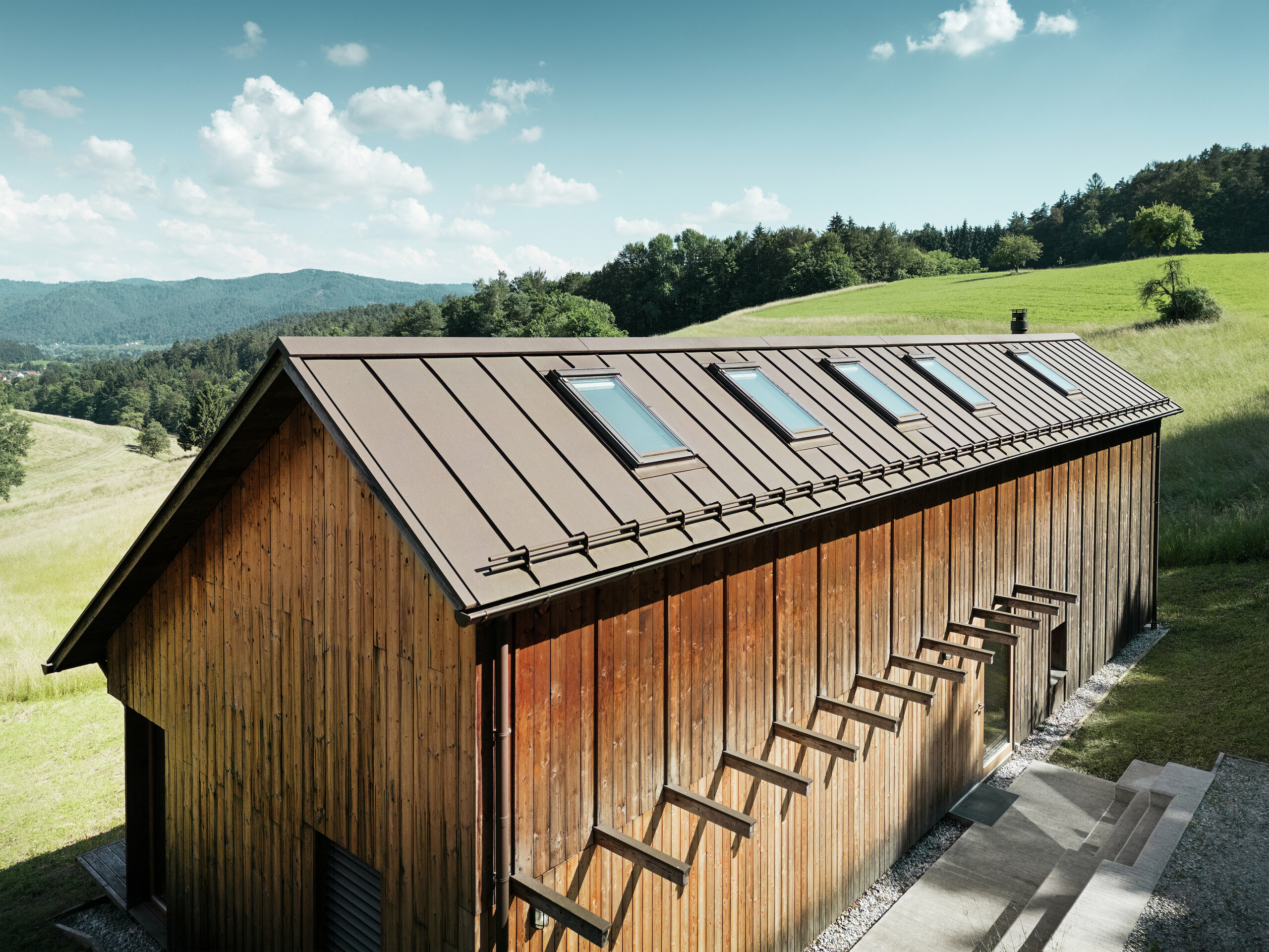 Architektonisch klar strukturiertes Holzhaus mit hochwertigem PREFALZ Aluminiumdach in P.10 Braun. Das wartungsarme PREFA Dach überzeugt durch exakte Linienführung, hohe UV- und Korrosionsbeständigkeit sowie perfekte Integration der Dachfenster. Die Kombination aus nachhaltiger Holzfassade und langlebiger Stehfalz-Dacheindeckung fügt sich harmonisch in die idyllische Hügellandschaft ein. Das asymmetrische Satteldach verfügt über fünf präzise verlegte Dachfenster, die für Licht im Inneren sorgen.