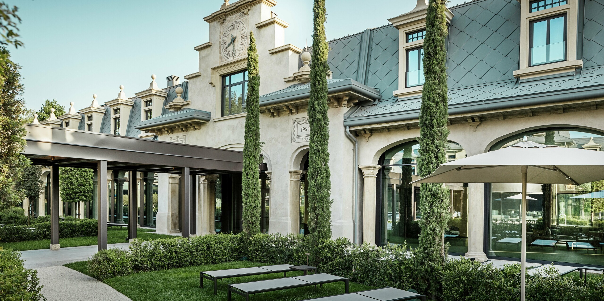 Detailreiche Außenansicht des Eingangbereichs zum urbanen Thermalpark „De Montel – Terme Milano“ im Mailänder Stadtteil San Siro. Die Fassade ist mit einer markanten Turmuhr und einem Baujahr-Relief mit der Zahl „1921“ versehen. Die Dacheindeckung mit PREFA Dachrauten 44 × 44 in P.10 Hellgrau unterstreicht die elegante Linienführung des Jugendstilensembles. Moderne Akzente, wie der schlanke Laubengang und die umliegende Gartenanlage mit Liegen und Zypressen, schaffen eine harmonische Verbindung aus Geschichte, Design und Funktion.