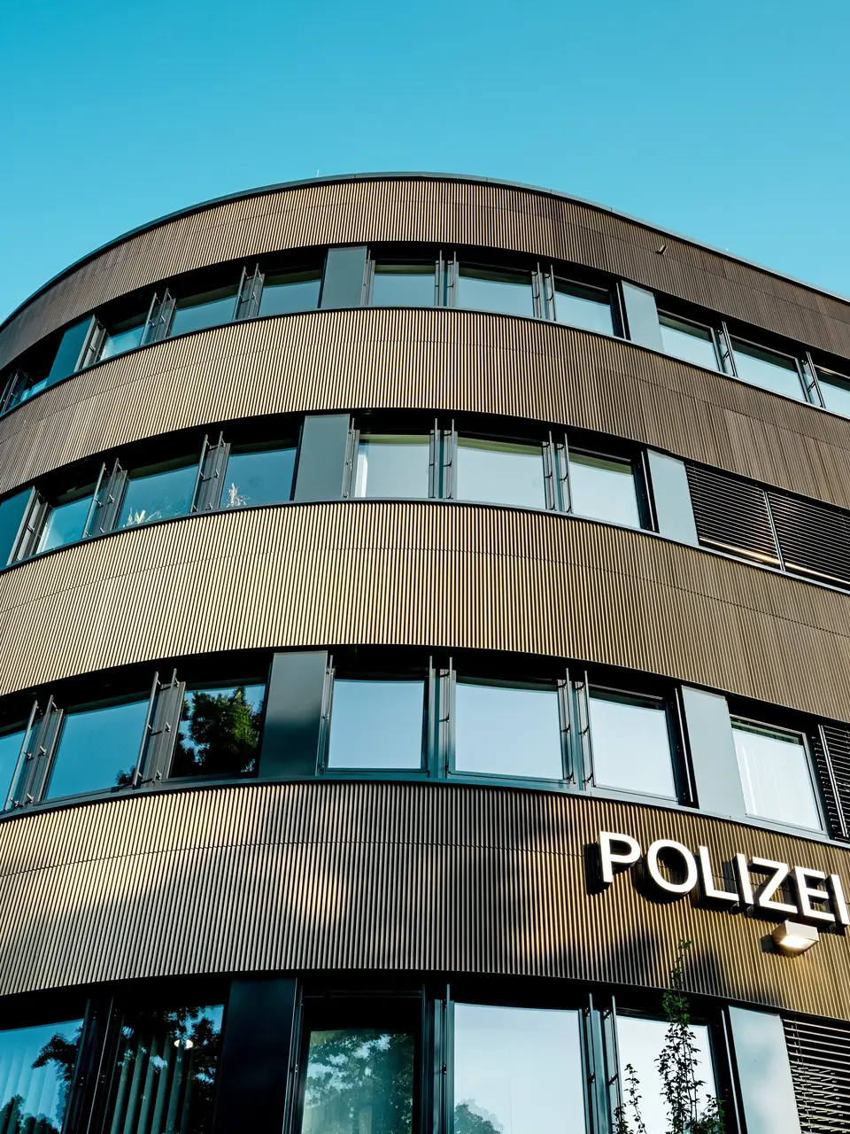 Dramatische Untersicht des geschwungenen Neubaus vom Polizeikommissariat 43 in Hamburg: Die markante PREFA Aluminiumfassade aus eloxierten Strangpressprofilen „Zacke“ in Mittelbronze zieht als architektonischer Eyecatcher alle Blicke auf sich. Die feine, rhythmische Struktur erzeugt dynamische Licht- und Schattenverläufe und betont die fließenden Rundungen des Baukörpers. PREFA Aluminium überzeugt durch hohe Witterungsbeständigkeit, Korrosionsschutz und Langlebigkeit – eine nachhaltige, wartungsarme Lösung für präzise, moderne Fassadenarchitektur.