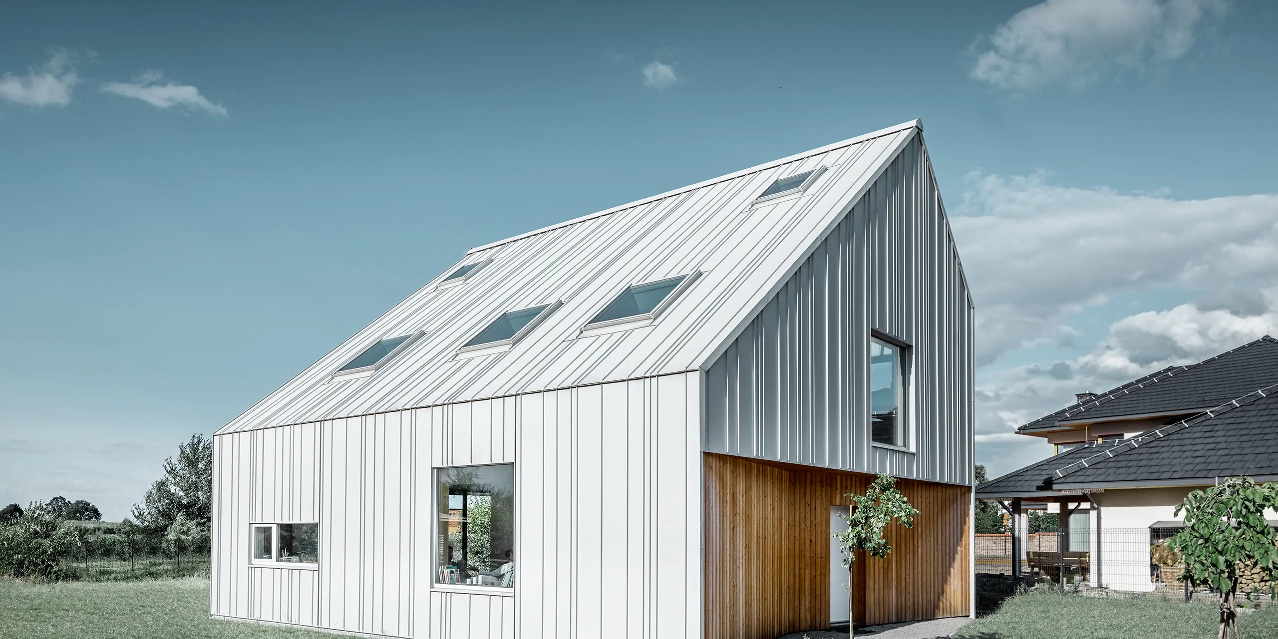 Modern detached house with gable roof, the entire outer skin is clad with PREFALZ standing seam in PREFA white. The panels of different widths extend from the roof over the façade. Several roof windows are installed in the roof.