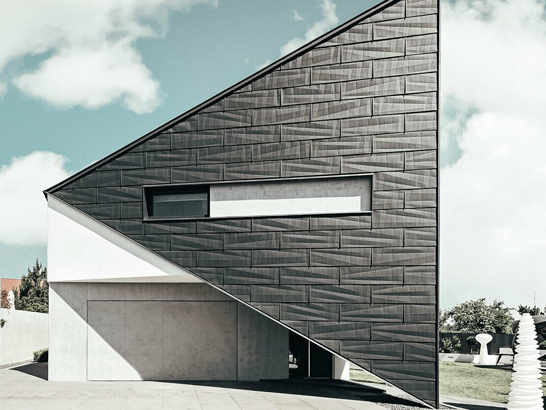 A front view of the triangle house in stettn, Poland. The façade is covered with the PREFA FX.12 panels in P.10 anthracite. It´s cloudy and a big window can be seen in the middle of the house.