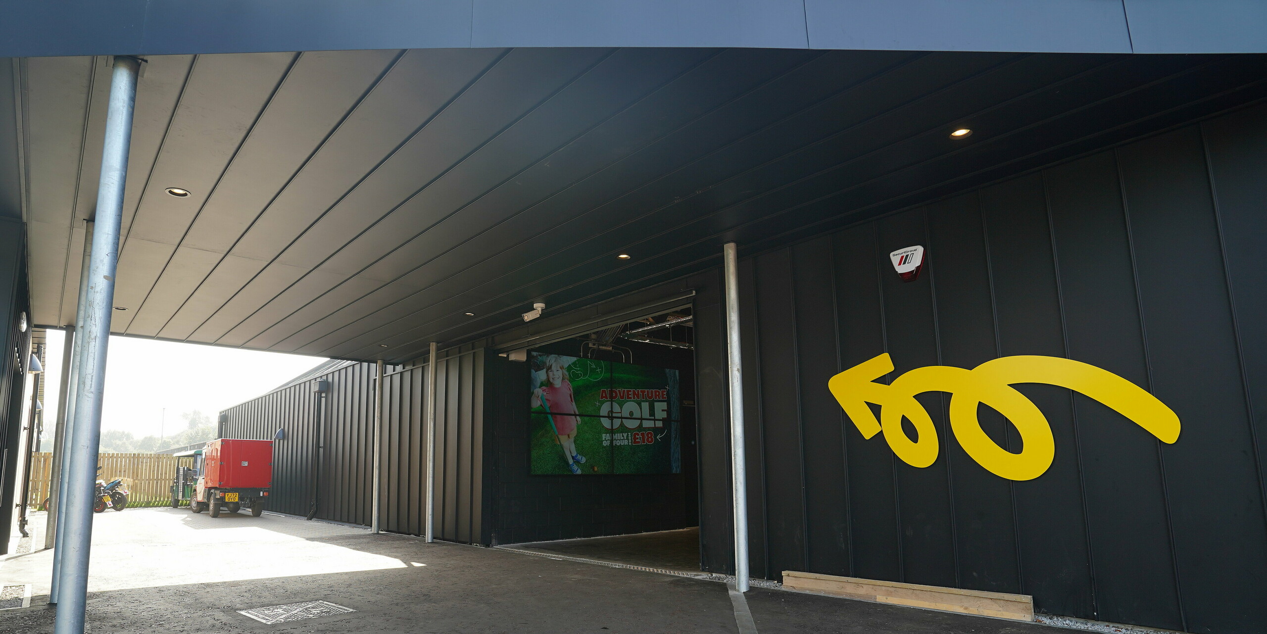 Entrance area of the 'Golf it! driving range and golf venue in Glasgow, Scotland, with a modern canopy made of PREFALZ in P.10 black. The aluminium elements create an inviting atmosphere, accentuated by a curved arrow in yellow on the wall and a large entrance sign. These details show how PREFALZ can be aesthetically and functionally integrated into different building areas.