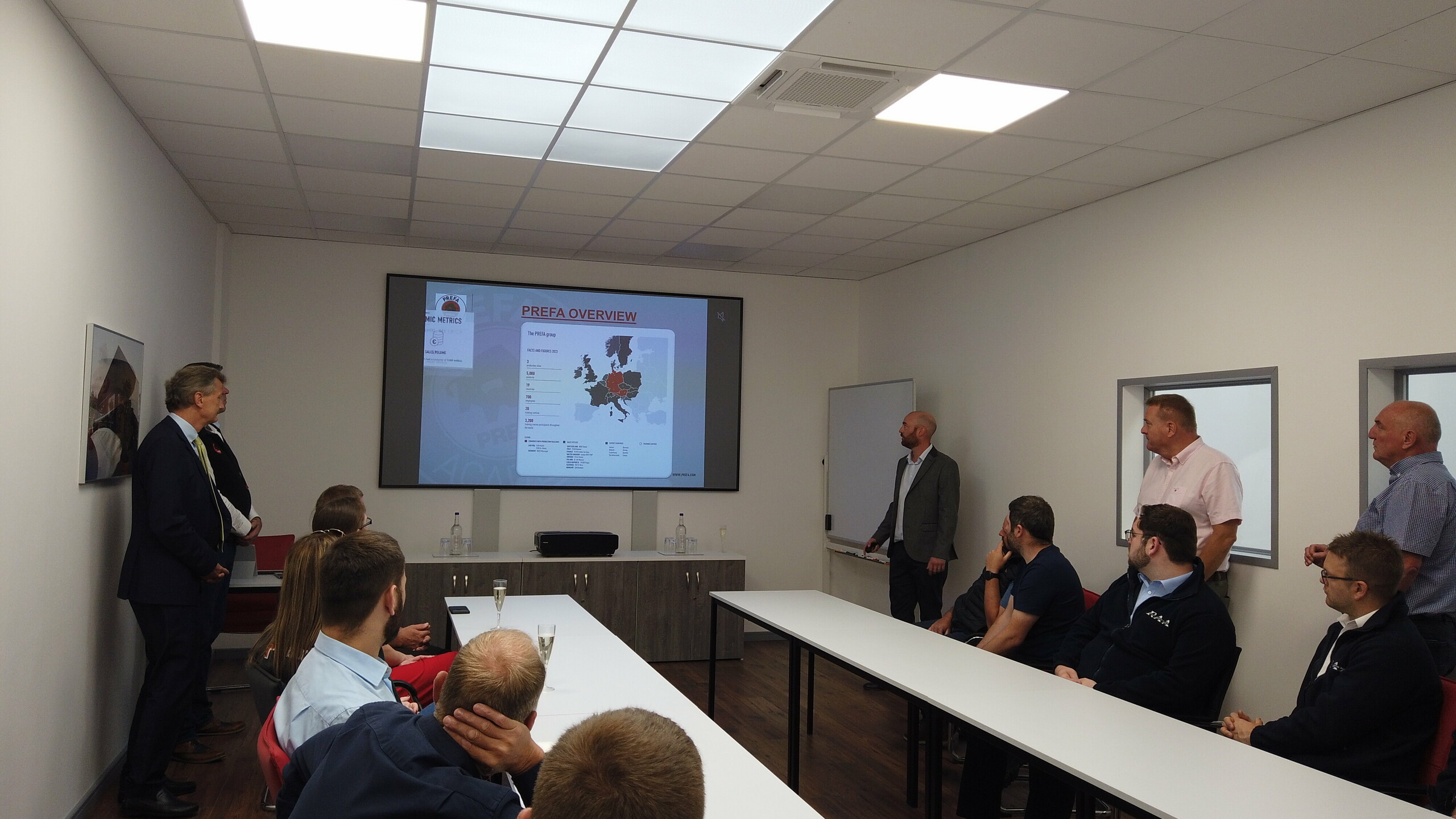 Photo from the opening presentation of the new Academy in the UK. Spectators sit at a table and watch the presentation.