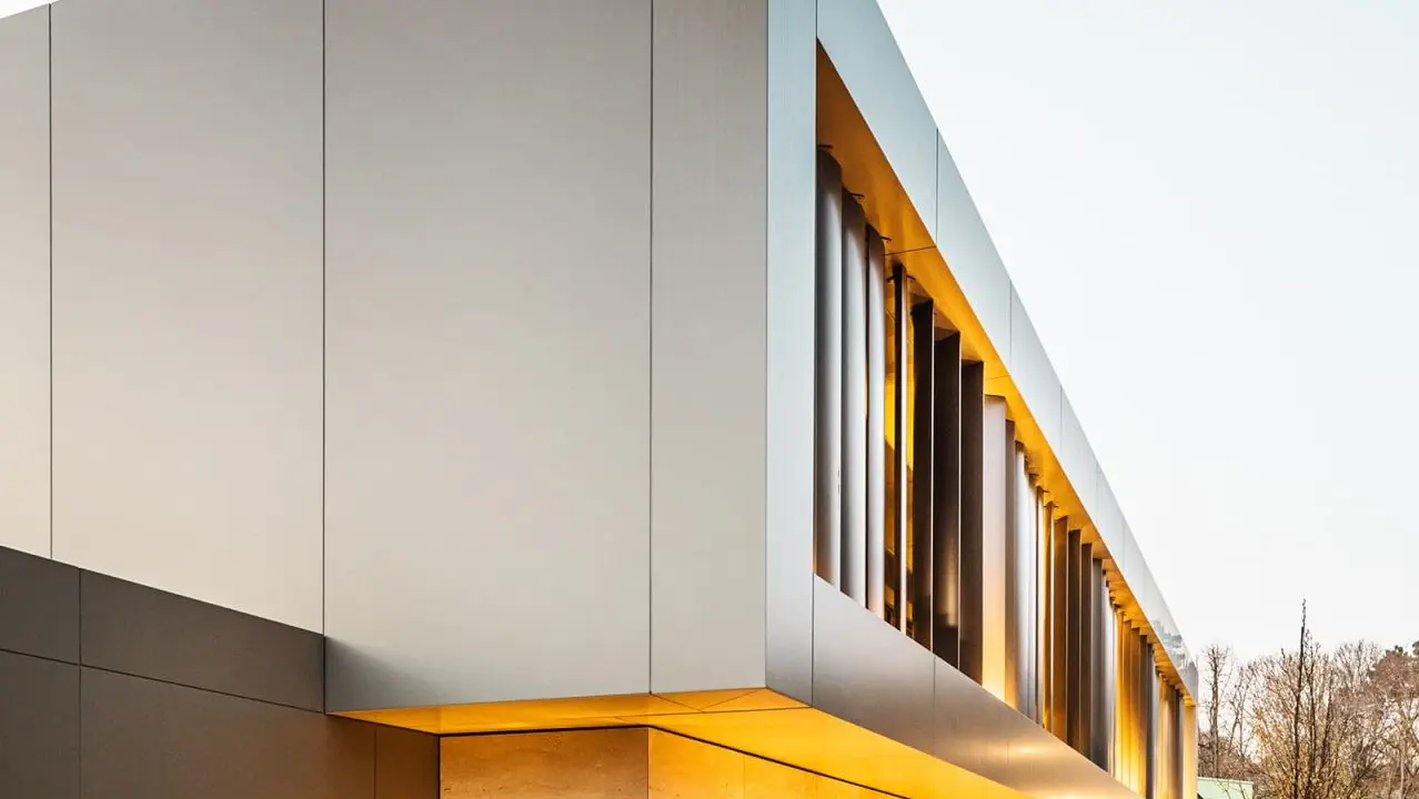 Diagonal view from the front – on the street side, bronze-coloured façade edge protrudes over light stone façade, a paved forecourt to the right of it