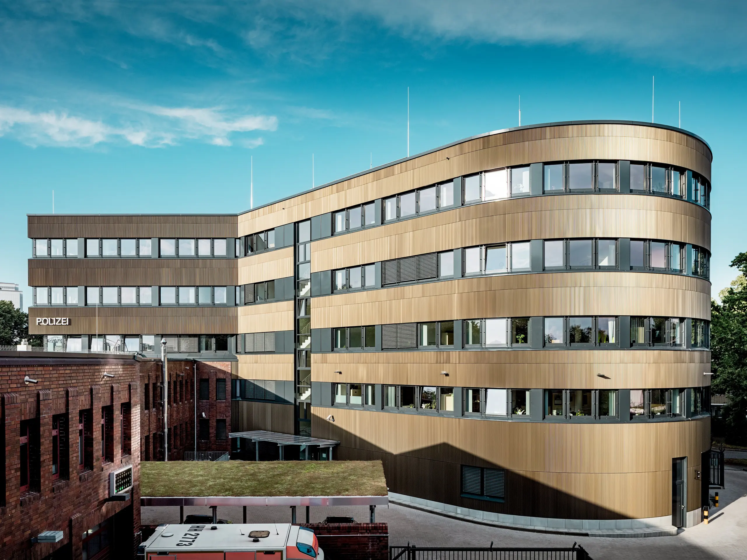 Markante Gesamtansicht des Polizeikommissariats 43 in Hamburg: Der geschwungene Neubau hebt sich mit seiner rund 1.100 m² großen PREFA Aluminiumfassade aus Strangpressprofilen „Zacke“ in Mittelbronze eindrucksvoll vom Backsteinbestand ab. Die fein strukturierte Oberfläche erzeugt lebendige Licht- und Schattenverläufe und macht das Gebäude zum architektonischen Eyecatcher. Dank hoher Witterungsbeständigkeit, Korrosionsschutz, Langlebigkeit und minimalem Wartungsaufwand stehen PREFA Aluminiumlösungen für nachhaltige, funktionale und designstarke Fassadenarchitektur.