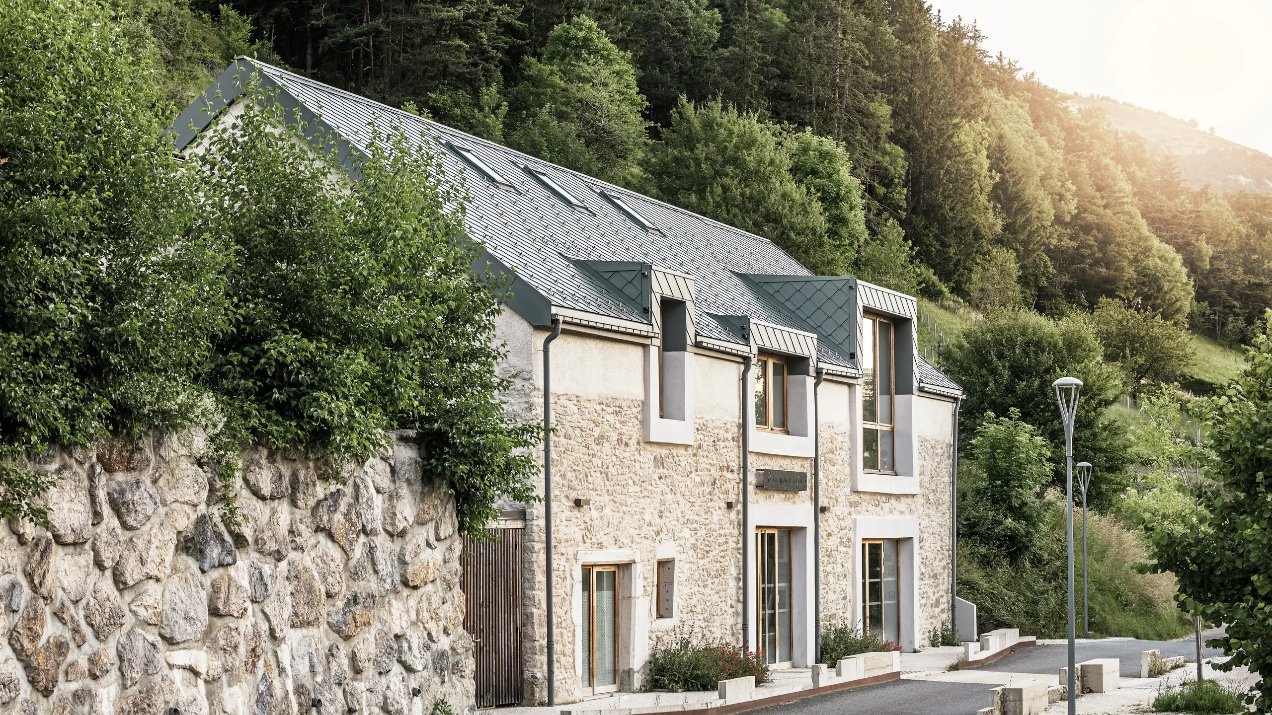 Side view of the restored barn ‘La Grange à Gaby’ in Engins, France. The building combines traditional stone masonry with a modern PREFA aluminium roof made of 29 × 29 cm rhomboid tiles. The careful renovation combines historical construction with long-lasting materials.