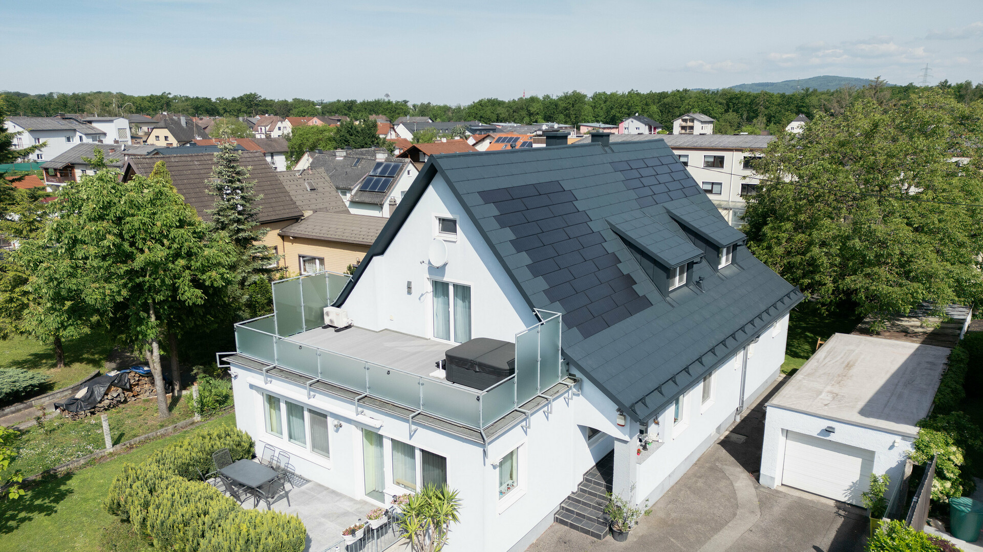 A bird’s-eye view of a detached house following roof renovation. The roof is now covered with large PREFA solar roof panels and PREFA R.16 roof tiles in P.10 anthracite. The solar roof panels are evenly distributed, creating a modern, sustainable solution. The building now has a modern and environmentally friendly appearance. The balcony in the foreground has a glass railing and is located on the upper floor of the building.