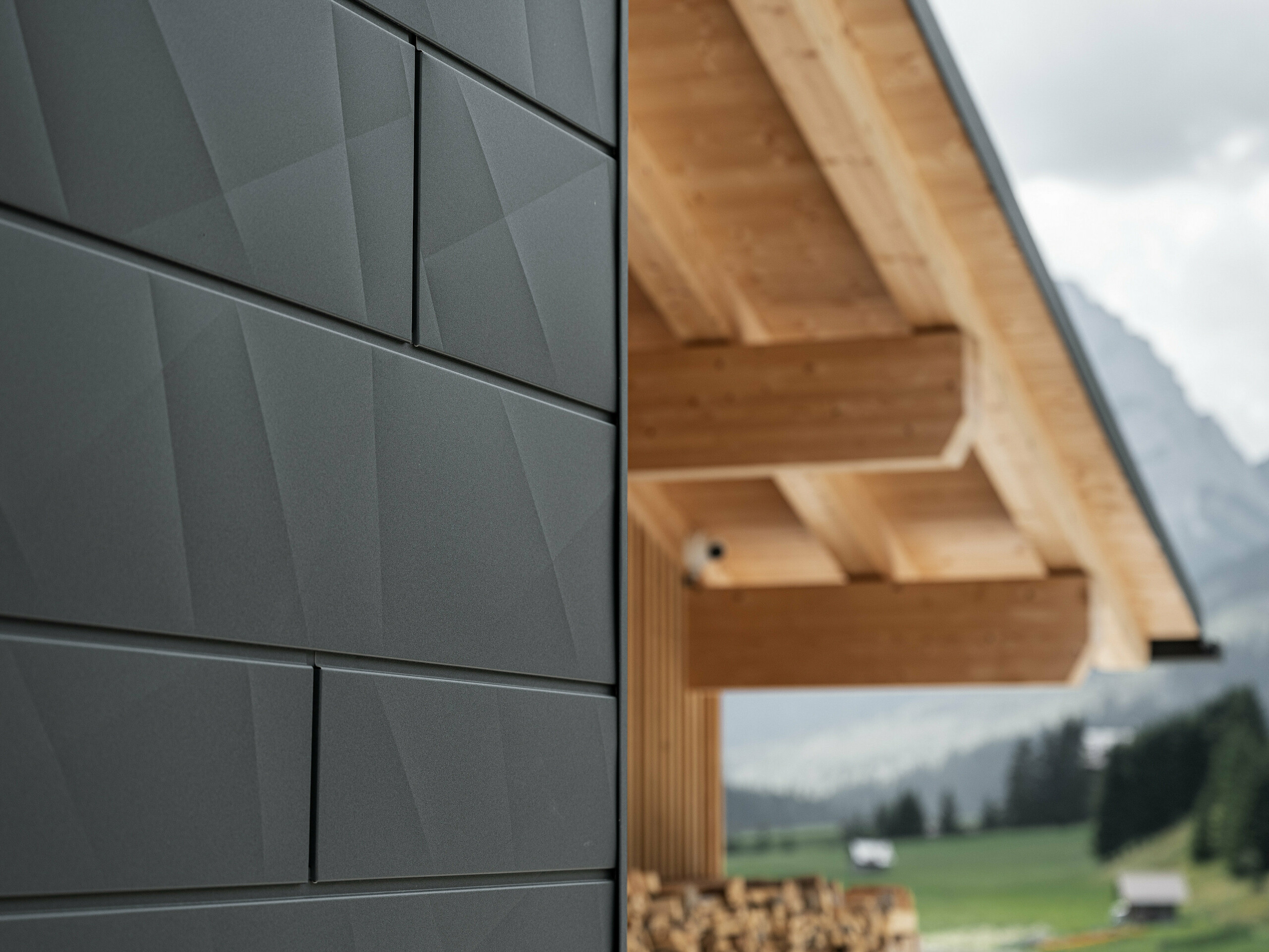 Close-up of PREFA Siding.X façade panels in P.10 black on Casa Hoffe in Sappada, Italy. The aluminium façade system is skillfully installed in a modern, abstract design, creating an appealing contrast with the visible wooden elements of the roof structure. The innovative façade solution blends seamlessly into the alpine setting and represents a combination of functionality, durability and aesthetic building culture.