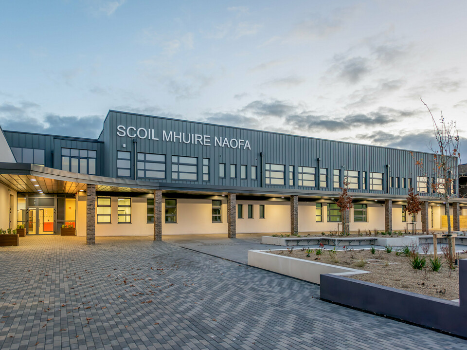 The Scoil Mhuire Naofa in Carrigtwohill, Ireland, at dusk. The contemporary façade made of PREFALZ aluminium in the colour P.10 light grey, combined with brick columns and clean lines, gives the school building an inviting and high-quality appearance. The clear architecture, stylish lighting and well-kept forecourt create a pleasant atmosphere.