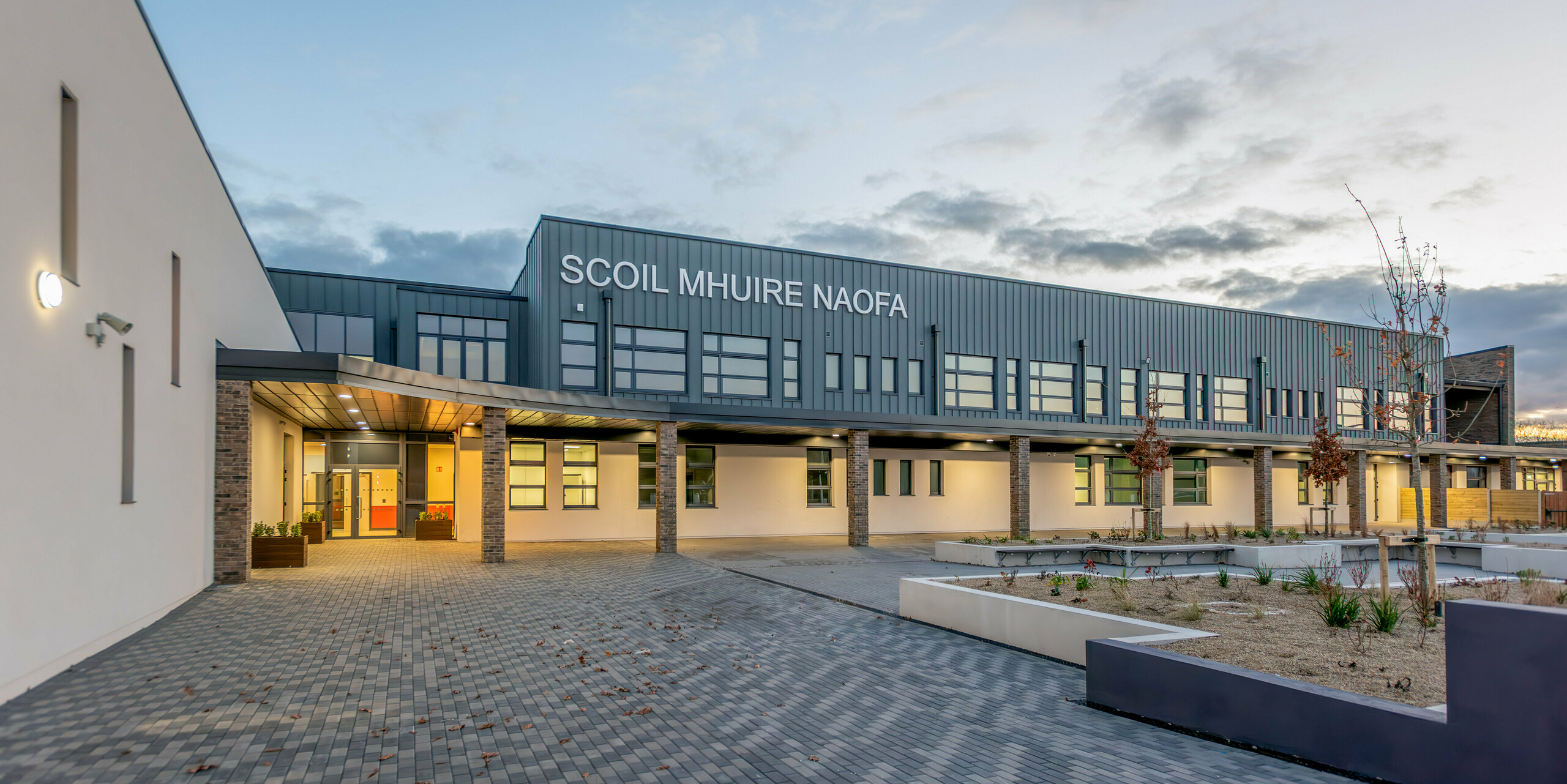 The Scoil Mhuire Naofa in Carrigtwohill, Ireland, at dusk. The contemporary façade made of PREFALZ aluminium in the colour P.10 light grey, combined with brick columns and clean lines, gives the school building an inviting and high-quality appearance. The clear architecture, stylish lighting and well-kept forecourt create a pleasant atmosphere.