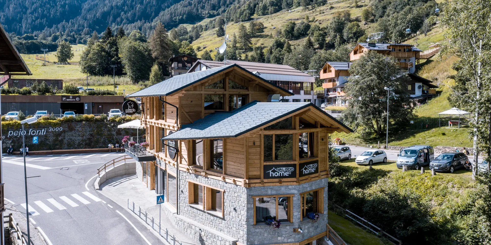Ansicht einer modernen Après-Ski-Bar in traditioneller Holzarchitektur mit steingrauem PREFA Dachschindeln DS.19, aus Aluminium integriert in die idyllische Berglandschaft mit üppigem Grün und Blick auf die Alpen.