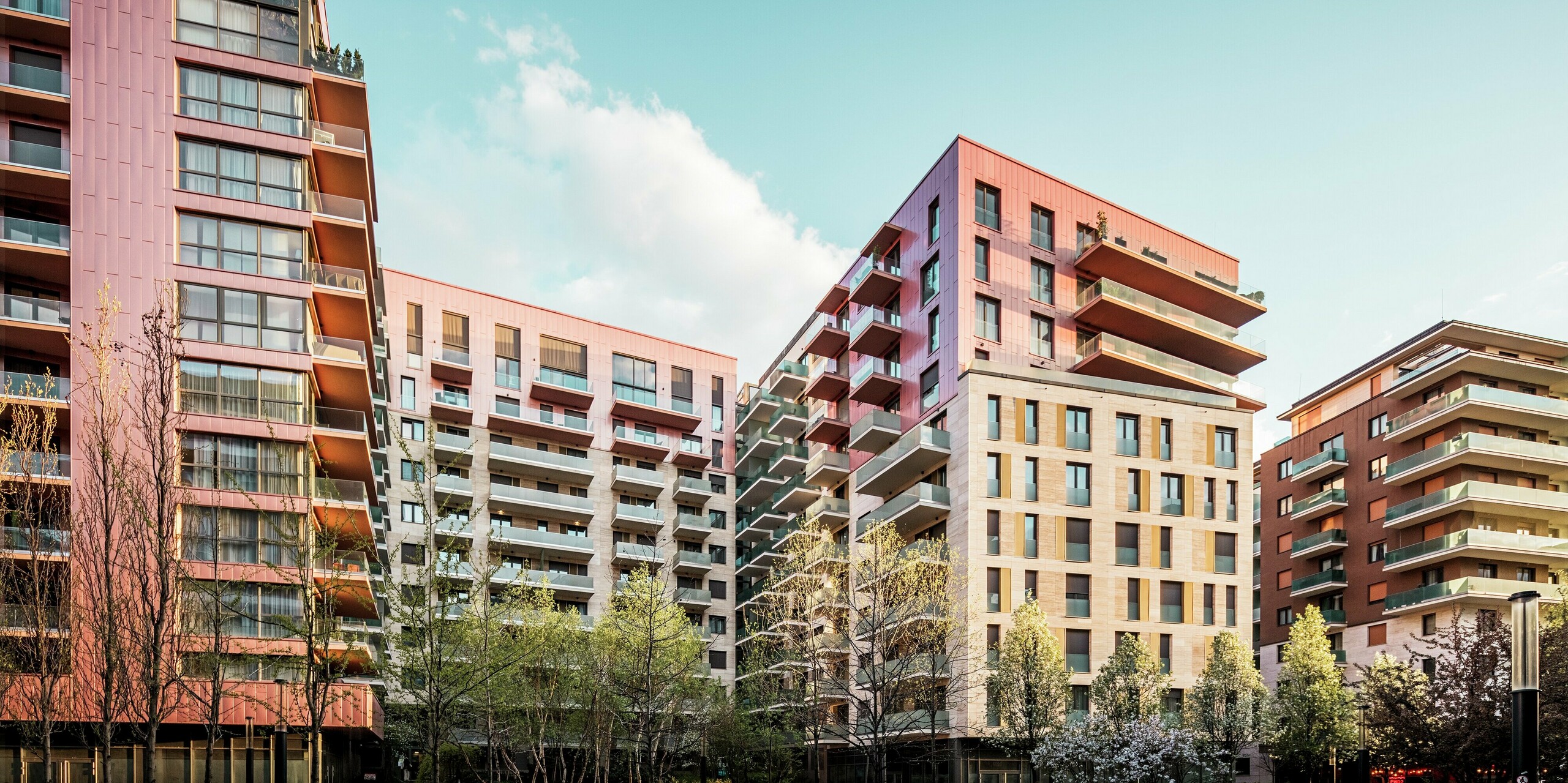 Gesamtansicht des Grand Corvin Wohnprojekts im revitalisierten Corvin-Viertel von Budapest. Die auffällige PREFA Siding Fassadenverkleidung in der Sonderfarbe Lachsmetallic setzt starke visuelle Akzente und betont die oberen Etagen. In Kombination mit Naturstein und Glas entsteht eine hochwertige, moderne Architektur. Die langlebigen Aluminiumpaneele sind pflegeleicht, witterungsbeständig und fügen sich harmonisch in das urbane Umfeld ein. Das Bild wurde während der Golden Hour aufgenommen, weshalb angenehmes, warmes Licht die Szene einrahmt.