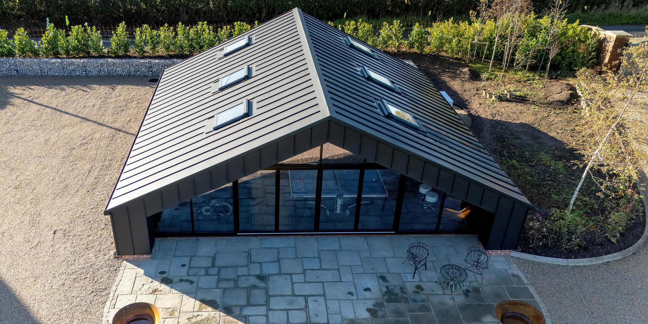 Aerial view of the modern leisure building clad entirely with PREFA aluminum and a PREFALZ standing seam roof in P.10 black. The symmetrical roof shape, expansive glass façade and defined outdoor patio create a striking architectural presence. The durable, weather‑resistant aluminum surface ensures long‑lasting stability and a premium design finish.