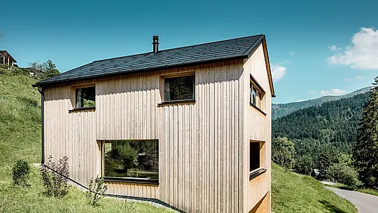 Sonnenlicht zeichnet markante Schatten auf die vertikale Weißtannenfassade von Haus Immi, das sich kompakt in den steilen Berghang von Ebnit einfügt. Über der klaren Holzarchitektur liegt das schwarze PREFA Solardach mit integrierten Solardachplatten und Dachplatten R.16. Die robuste Aluminiumlösung erzeugt Strom, schützt dauerhaft und bewahrt die reduzierte Gebäudeform optisch unverändert.