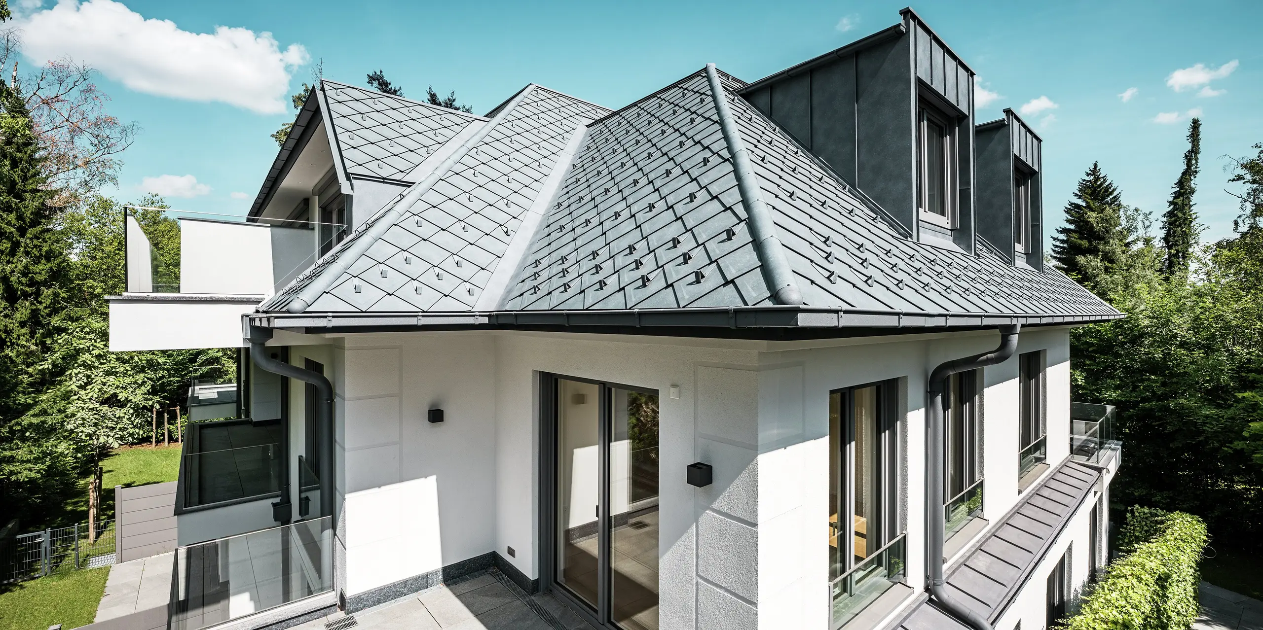 Modernes Mehrfamilienhaus mit markanter Dacharchitektur in PREFA P.10 Steingrau: Die Dachfläche ist mit hochwertigen PREFA Dachrauten 29 × 29 gedeckt und sorgt für ein edles, strukturiertes Erscheinungsbild. Stilvolle Flachdachgauben aus PREFALZ fügen sich nahtlos ins Design ein. Integrierte Schneestopper und eine formschöne PREFA Dachentwässerung mit Kastenrinne und Ablaufrohr unterstreichen die technische Raffinesse. Das langlebige Aluminiumdach trotzt jeder Witterung und vereint Ästhetik mit Funktionalität – ideal für zeitgemäßen Wohnbau.