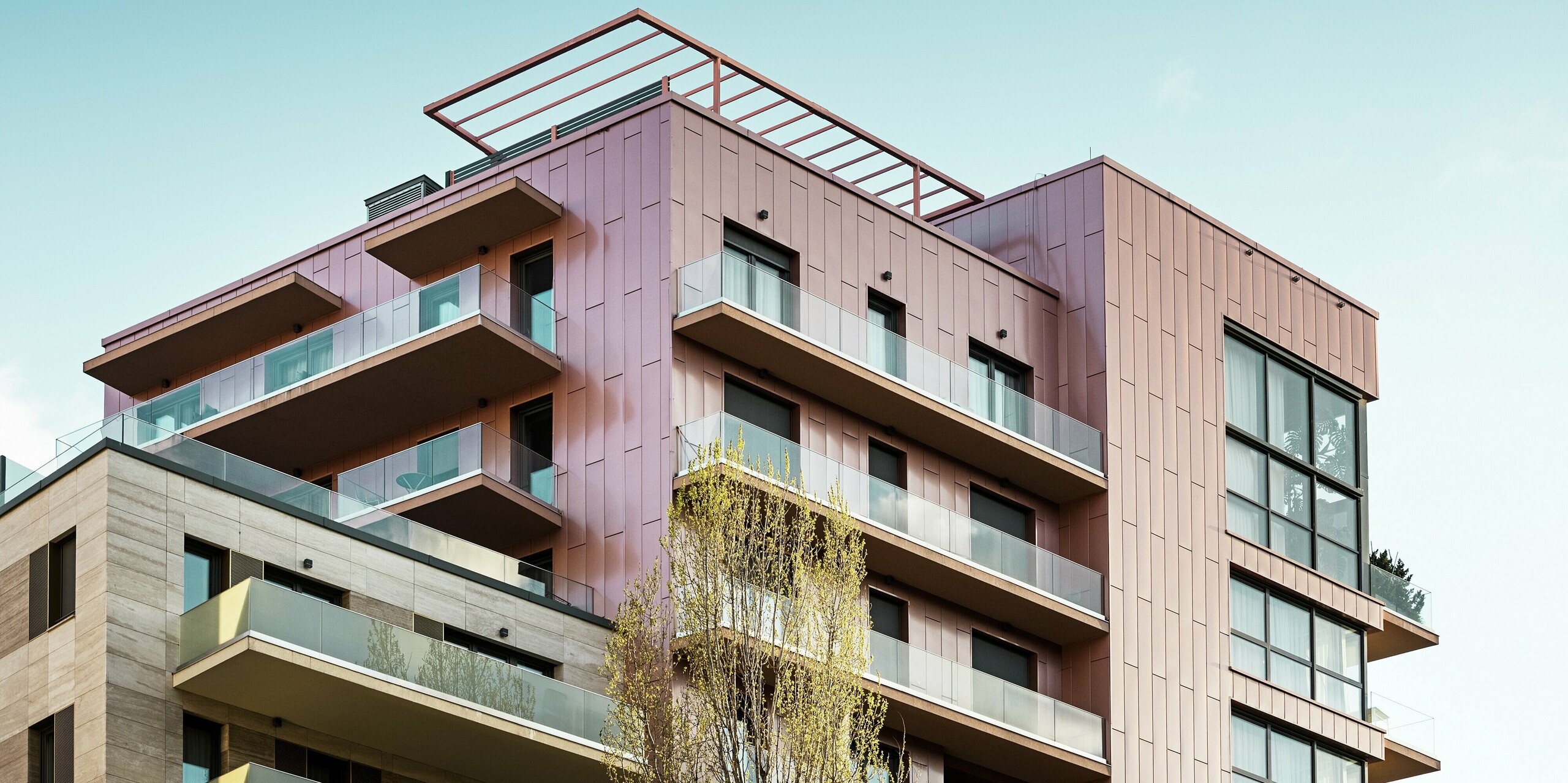 Oberer Gebäudeteil des Grand Corvin Wohnkomplexes mit großzügigen Glasflächen und modern gestalteten Balkonen, eingerahmt von vertikal verlegten PREFA Siding Fassadenpaneelen in der Sonderfarbe Lachsmetallic. Die langlebige, witterungsbeständige Aluminiumverkleidung unterstreicht das architektonische Konzept aus hochwertiger Materialität und urbanem Design im Herzen von Budapest.