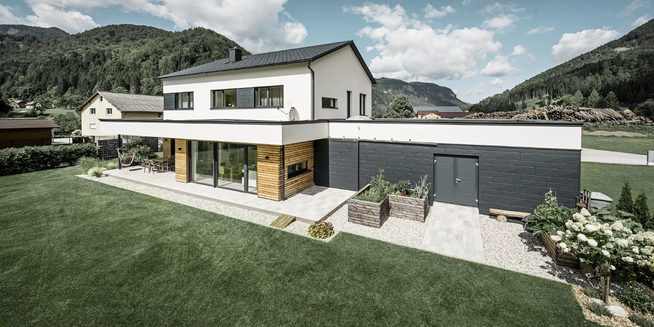 Modern single-family house with white rendered façade and PREFA Siding.X in P.10 anthracite, which forms a striking contrast to the natural environment. Covered with PREFA roof tiles, roof drainage with PREFA half-round gutter and downpipe.