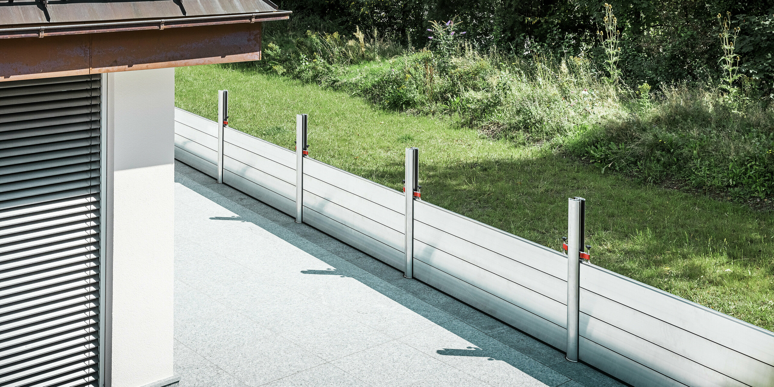 Schrägansicht des installierten PREFA Hochwasserschutzsystems entlang der Terrasse eines Wohnhauses in Tirol. Die robusten Dammbalken aus Aluminium sind zwischen fix verankerten Stützprofilen montiert, die mit Bodenhülsen fest im Untergrund verankert sind. Der mobile Hochwasserschutz bietet einen zuverlässigen Schutz vor möglichen Überflutungen aus dem angrenzenden Grünbereich. Im Vordergrund ist die gegliederte Fassade mit Jalousien zu sehen, während sich das Schutzsystem dezent und funktional in das hochwertige Wohnumfeld einfügt.