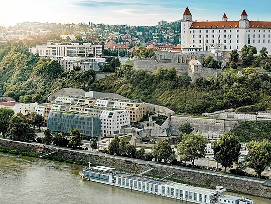 Weitblick über das Vydrica‑Quartier in Bratislava mit Blick auf die Burg und die Donau: Die vielschichtige Architektur fasziniert mit begrünten Dachflächen, steilen Satteldächern und präzise integrierten Dachfenstern als visuelle Highlights. PREFA Aluminium Dachschindeln in Prefaweiß, Sandbraun und Bronze verbinden die Baukörper zu einem harmonischen Gesamtbild und fügen sich in das historische Umfeld ein. Die langlebigen, leichten und extrem witterungsbeständigen PREFA Lösungen ermöglichen komplexe Dachgeometrien und sorgen für eine nachhaltige, wartungsarme Gebäudehülle mit hoher gestalterischer Qualität.