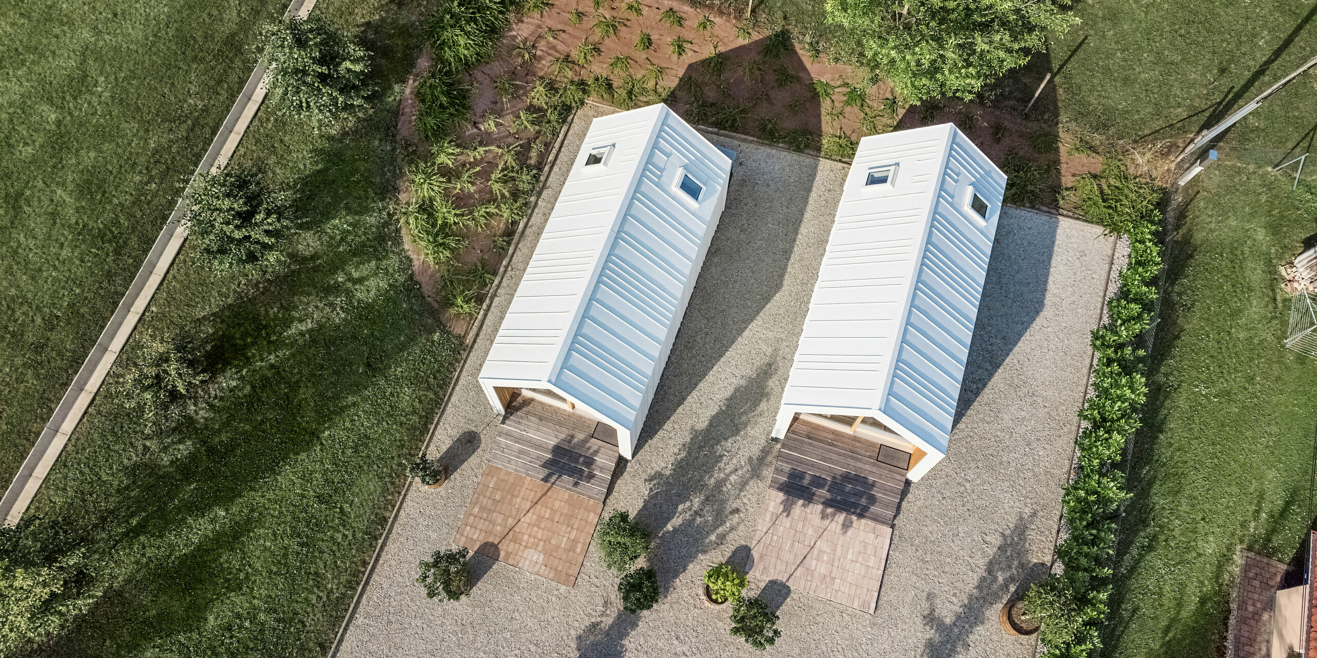 Bird's eye view of the two Tiny Houses at the 'Pri Momi' holiday complex in Kančevci, Slovenia, which are clad with PREFALZ roof and façade cladding in P.10 Pure White. The picture shows the symmetrical arrangement of the two buildings with their white cladding made of high-quality, robust sheet metal and the surrounding green areas. The clear lines and modern architecture of the buildings are emphasised by the precise design of the aluminium cladding. The surrounding paths and natural surroundings blend harmoniously into the overall picture and invite guests to stay a while.