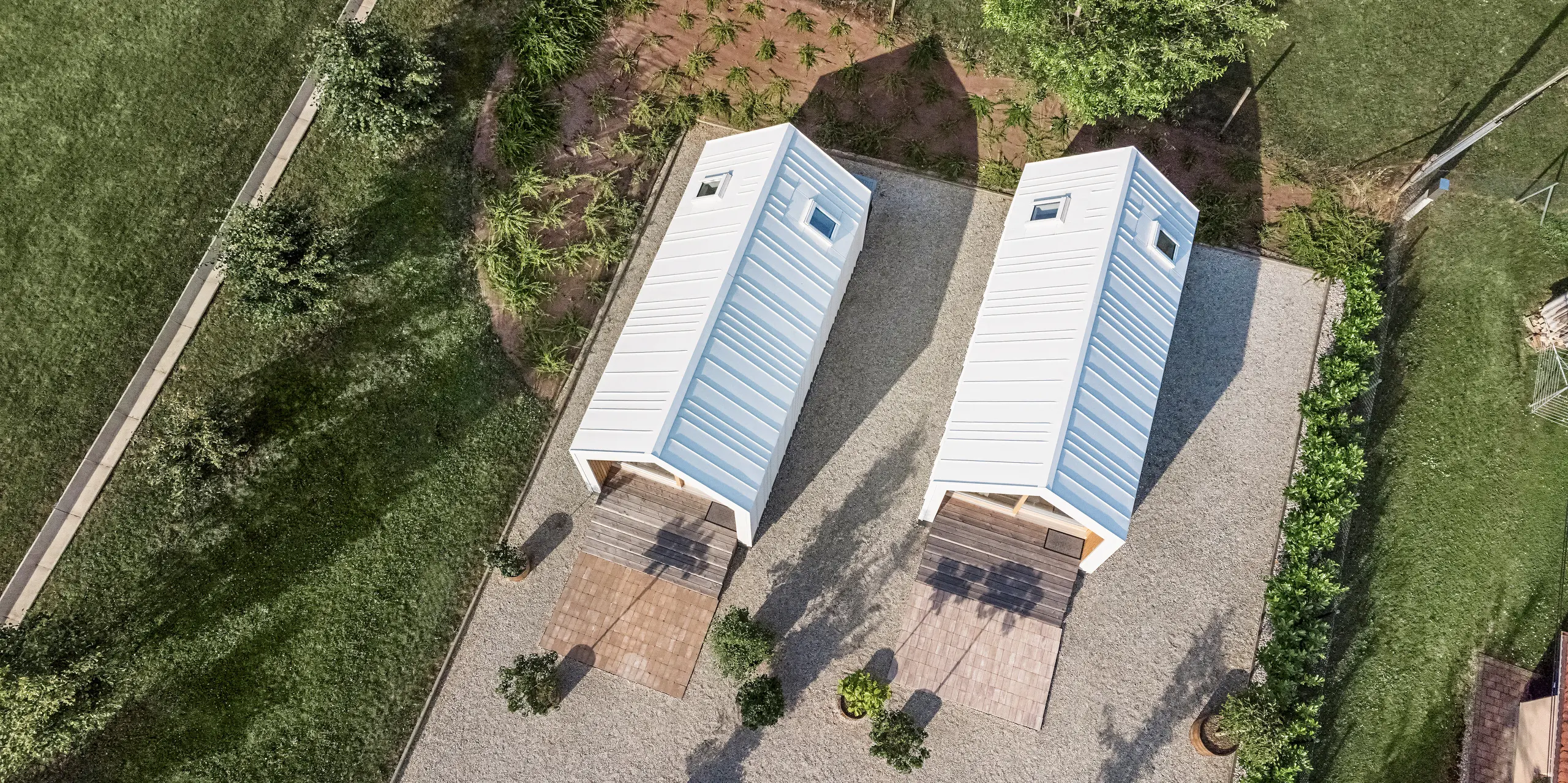 Bird's eye view of the two Tiny Houses at the 'Pri Momi' holiday complex in Kančevci, Slovenia, which are clad with PREFALZ roof and façade cladding in P.10 Pure White. The picture shows the symmetrical arrangement of the two buildings with their white cladding made of high-quality, robust sheet metal and the surrounding green areas. The clear lines and modern architecture of the buildings are emphasised by the precise design of the aluminium cladding. The surrounding paths and natural surroundings blend harmoniously into the overall picture and invite guests to stay a while.