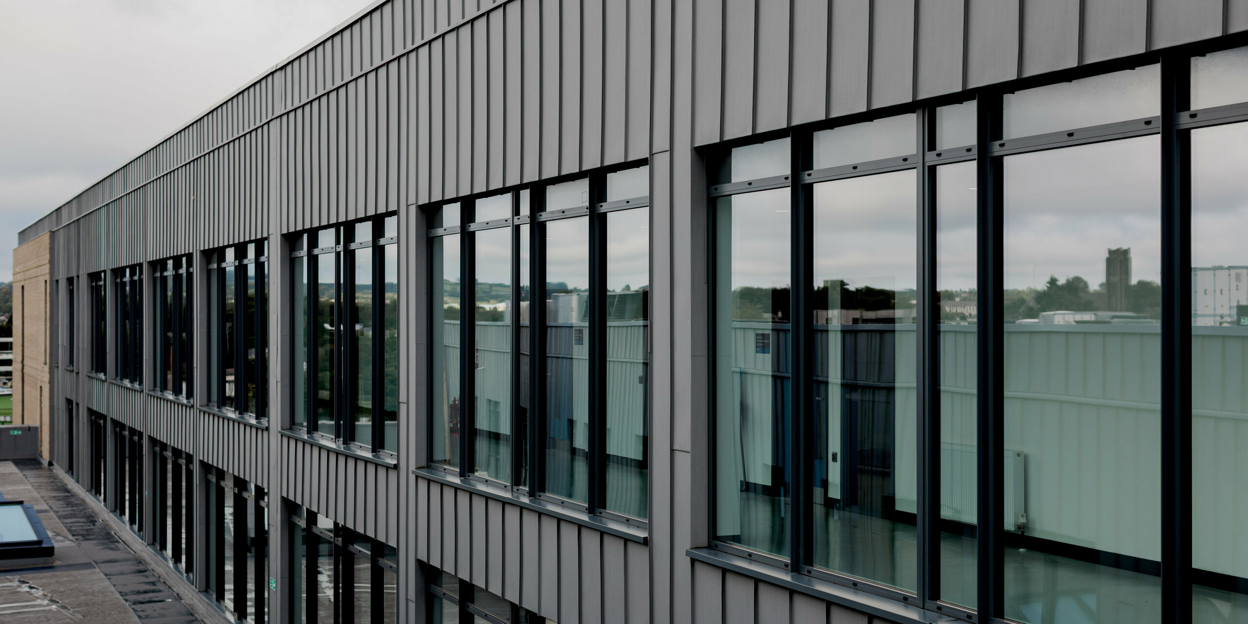 A long façade view of the Northern Regional College in Ballymena reveals a continuous PREFALZ aluminium cladding in patina grey with a strong vertical standing seam rhythm. Large, evenly spaced windows reflect the surrounding landscape and sky, adding depth and transparency to the elevation. The calm metallic surface frames the glazing precisely and enhances the building’s linear architectural language. PREFA aluminium delivers durability, weather resistance and a refined, low-maintenance façade solution for contemporary educational architecture.