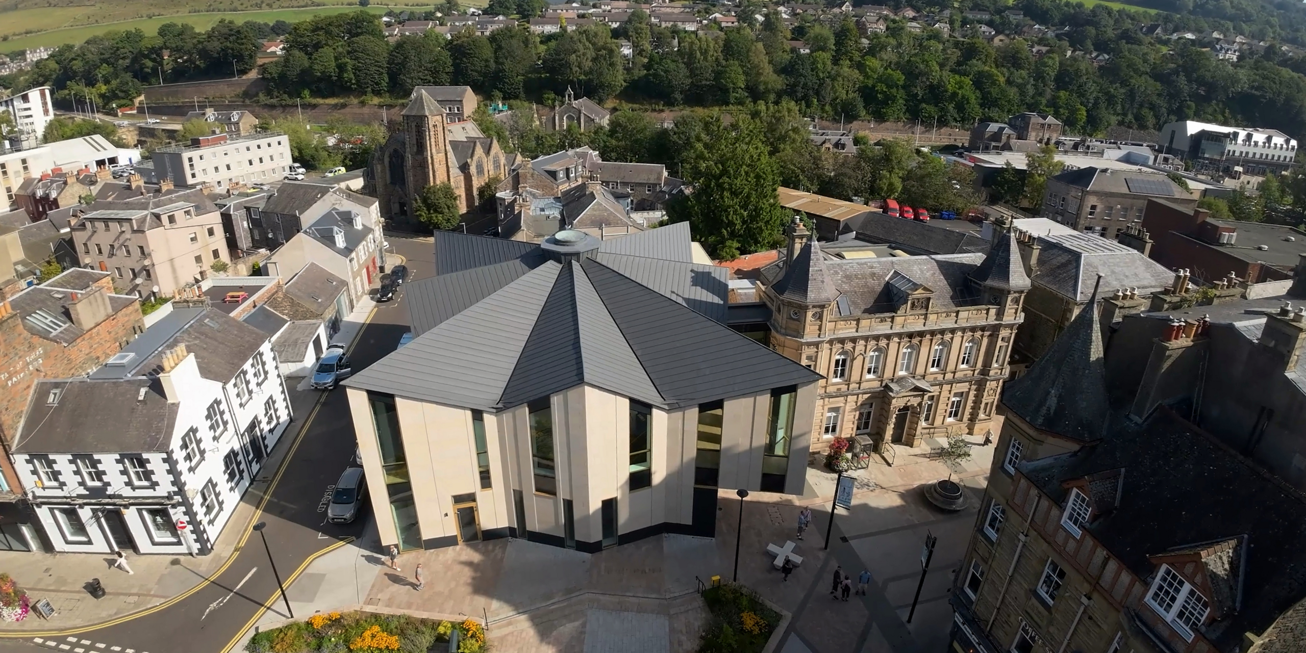 Drone shot of the visitor centre 'The Great Tapestry of Scotland'. The historic building is highlighted by a special roof landscape clad with PREFALZ in P.10 Zinc Grey. The unique roof surface with the contemporary and durable aluminium cladding blends harmoniously into the old town of Galashiels. The metallic colour scheme and clean lines of the roof reflect the cultural commitment of the centre, while at the same time blending respectfully into the historic townscape.
