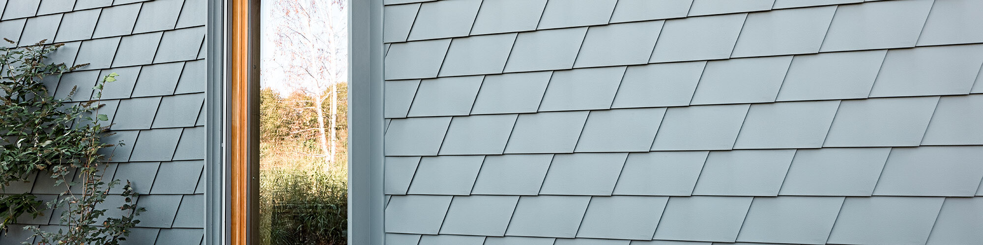 Detail view of the PREFA façade, a window reflects the natural surroundings, greenery grows up the façade on the left in the picture.