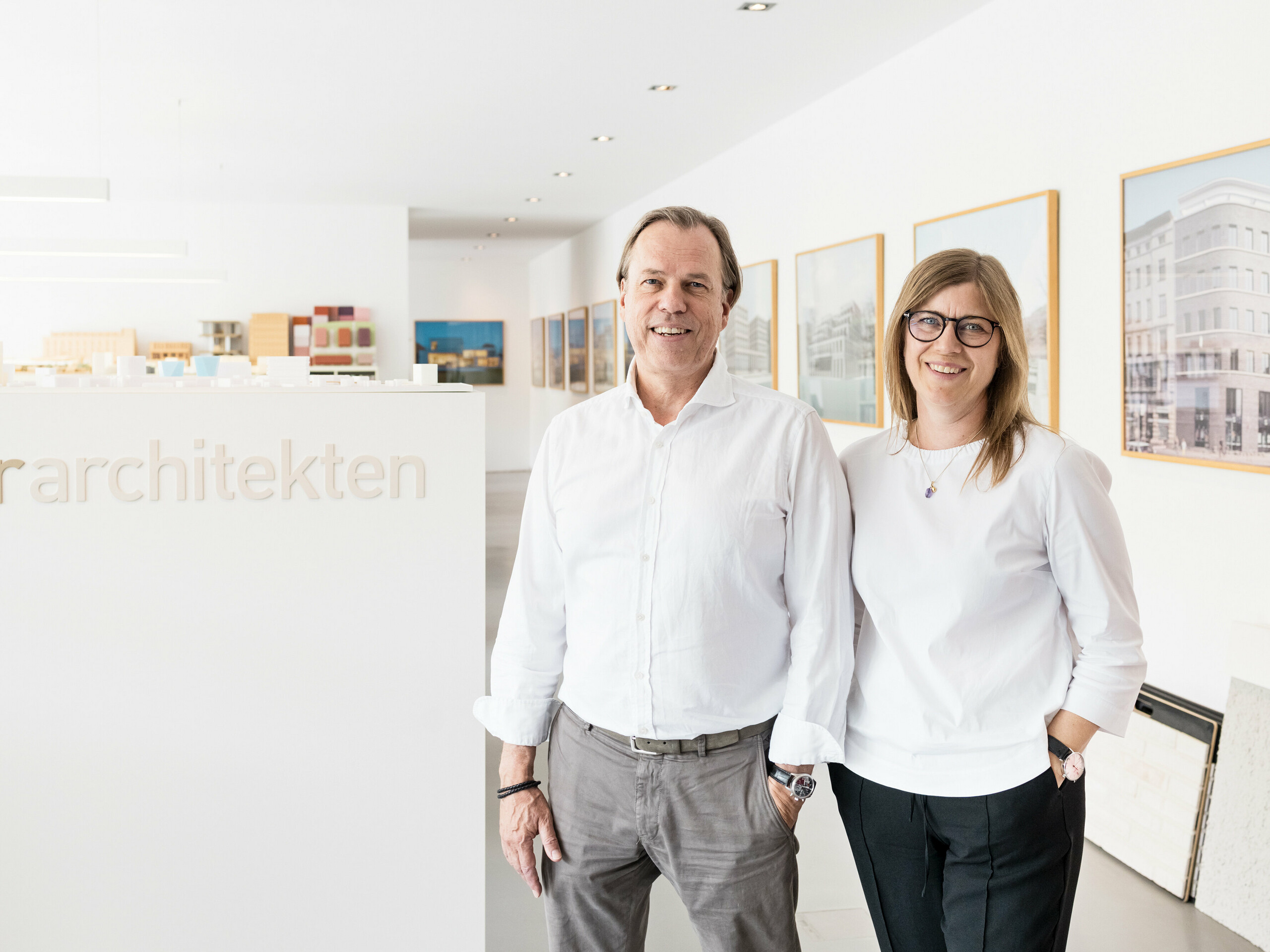 Leona and Andreas Geitner, the lead architects of the project in Ratingen (DE), stand smiling in front of a white wall with the lettering ‘Geitner Architekten’. Both are wearing white shirts, which gives the picture a professional and uniform appearance. Architectural models and framed pictures of building projects can be seen in the background, reflecting the creative and innovative environment of the architecture firm. The friendly and inviting atmosphere emphasises the professionalism and expertise of Leona and Andreas Geitner's team.