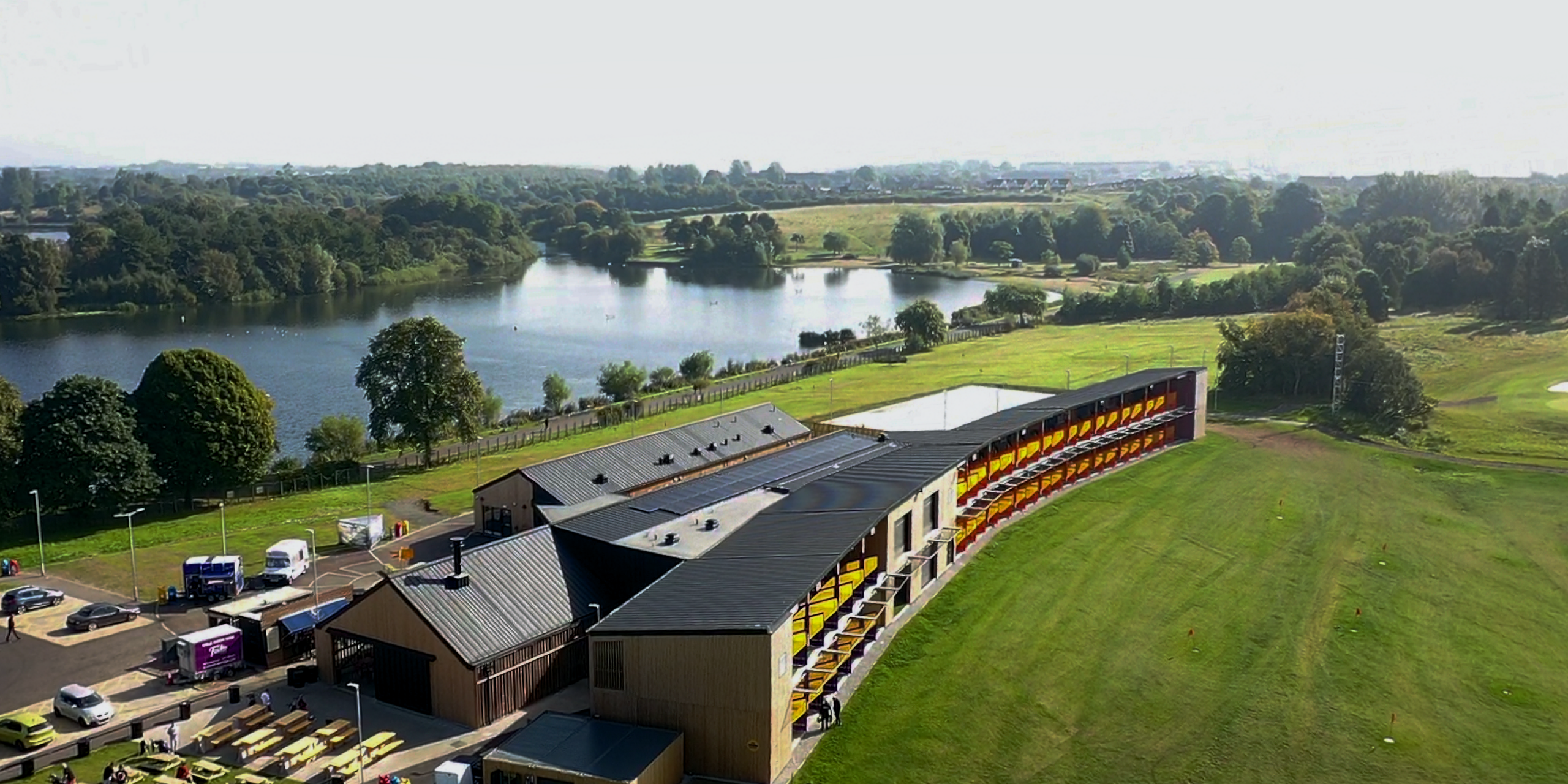 Drone shot of the 'Golf it!' driving range and golf venue in Glasgow, Scotland, with a PREFALZ roof system in P.10 black. The dark roof design creates an elegant contrast to the wooden façade of the building complex. The structure blends seamlessly into the surrounding landscape, with a clear view of the neighbouring lake and the lush greenery of the golf course. The roofs of the individual building components with their uniform, black Prefa aluminium cladding stand out against the natural backdrop and demonstrate the harmonious combination of modern architecture and natural beauty.