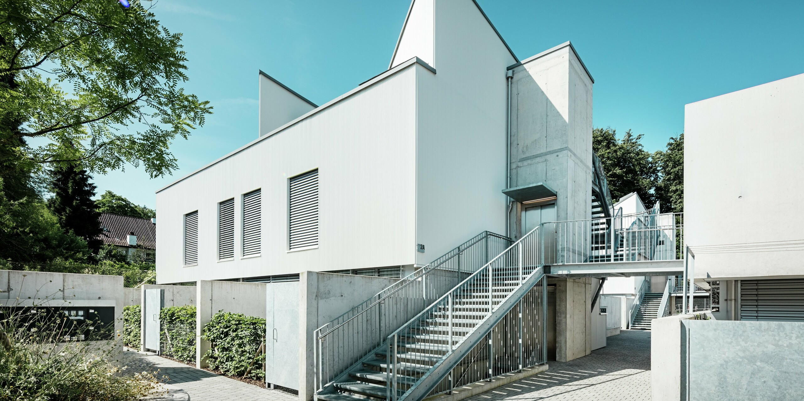 Ensemble moderner Tiny Homes mit markanten Spitzdächern, verkleidet mit dem PREFA Zackenprofil in der Farbe P.10 Reinweiß. Die schlichte, klare Fassadengestaltung harmoniert mit den Sichtbetonelementen und der umlaufenden Stahlkonstruktion. Die langlebige PREFA Aluminiumfassade schützt zuverlässig vor Witterung und sorgt für eine wartungsarme, ästhetisch reduzierte Optik.