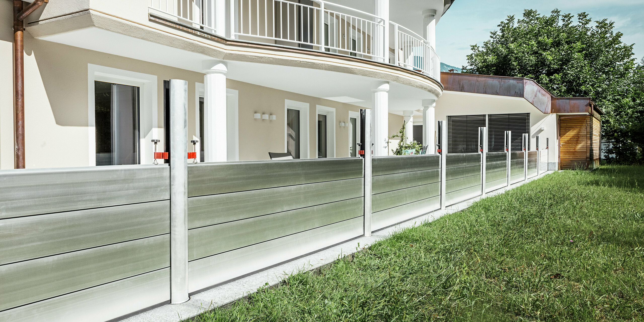 Panoramaansicht eines exklusiven Wohnhauses in Tirol mit installiertem PREFA Hochwasserschutzsystem entlang der Terrasse. Die Dammbalken aus Aluminium verlaufen über die gesamte Gebäudelänge und schützen mehrere Zugänge wie Terrassentüren und Fenster vor möglichem Hochwasser aus dem angrenzenden Gelände. Die moderne Architektur des Hauses mit Rundbalkon und weißen Stützen bildet einen stilvollen Kontrast zum funktionalen Hochwasserschutz aus Aluminium.