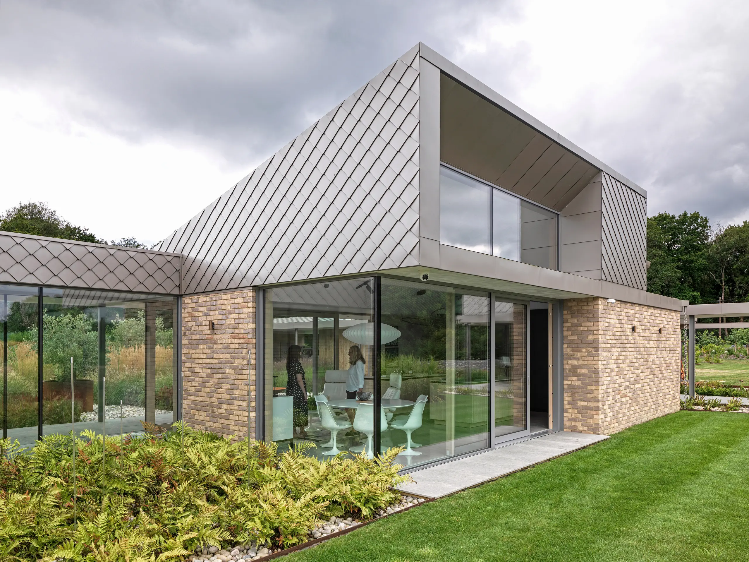 Architecturally striking part of a residential building in West Sussex with a modern façade design featuring PREFA rhomboid façade tiles 20 × 20 in P.10 bronze. The roof surfaces are covered with standing seam and their clear lines emphasise the timeless character of the design. Generous glass fronts create bright living spaces and visually connect the building with the well-kept garden. The high-quality PREFA products offer durability and a uniform appearance with minimal maintenance.