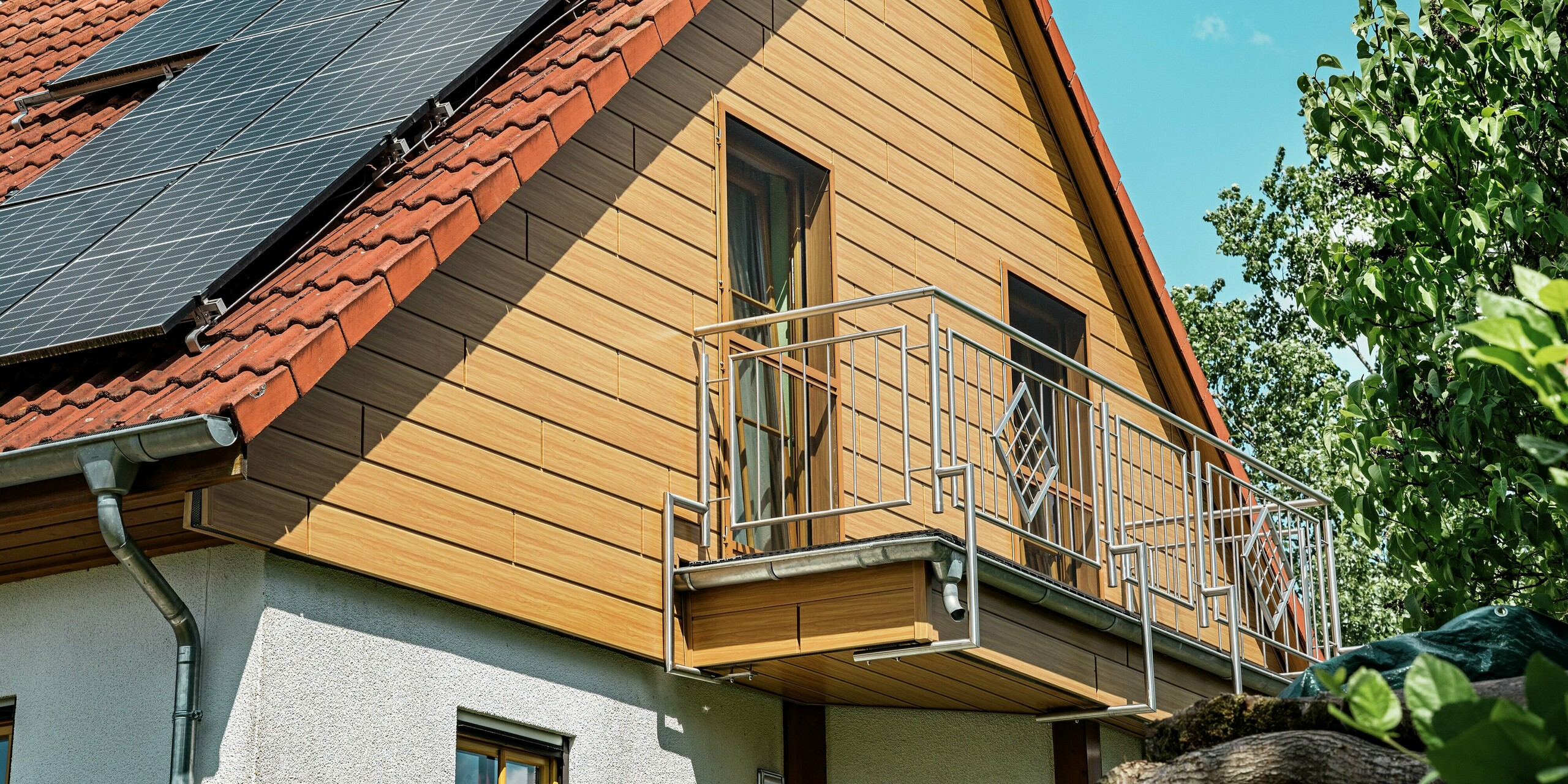 Das Bild zeigt den frisch sanierten Giebel eines Einfamilienhauses in Blieskastel, der mit PREFA Sidings in Eiche Natur verkleidet wurde. Die warme Holzoptik aus langlebigem Aluminium harmoniert perfekt mit dem roten Ziegeldach und der großen PV-Anlage, die imposant über die Dachfläche verläuft. Der filigrane Balkon aus Metall unterstreicht die moderne Anmutung und bildet einen spannenden Kontrast zur natürlichen Fassadenstruktur. Dank der witterungsbeständigen, farbstabilen PREFA Oberfläche bleibt dieser Look dauerhaft erhalten – ein echtes Highlight für das gesamte Gebäude.