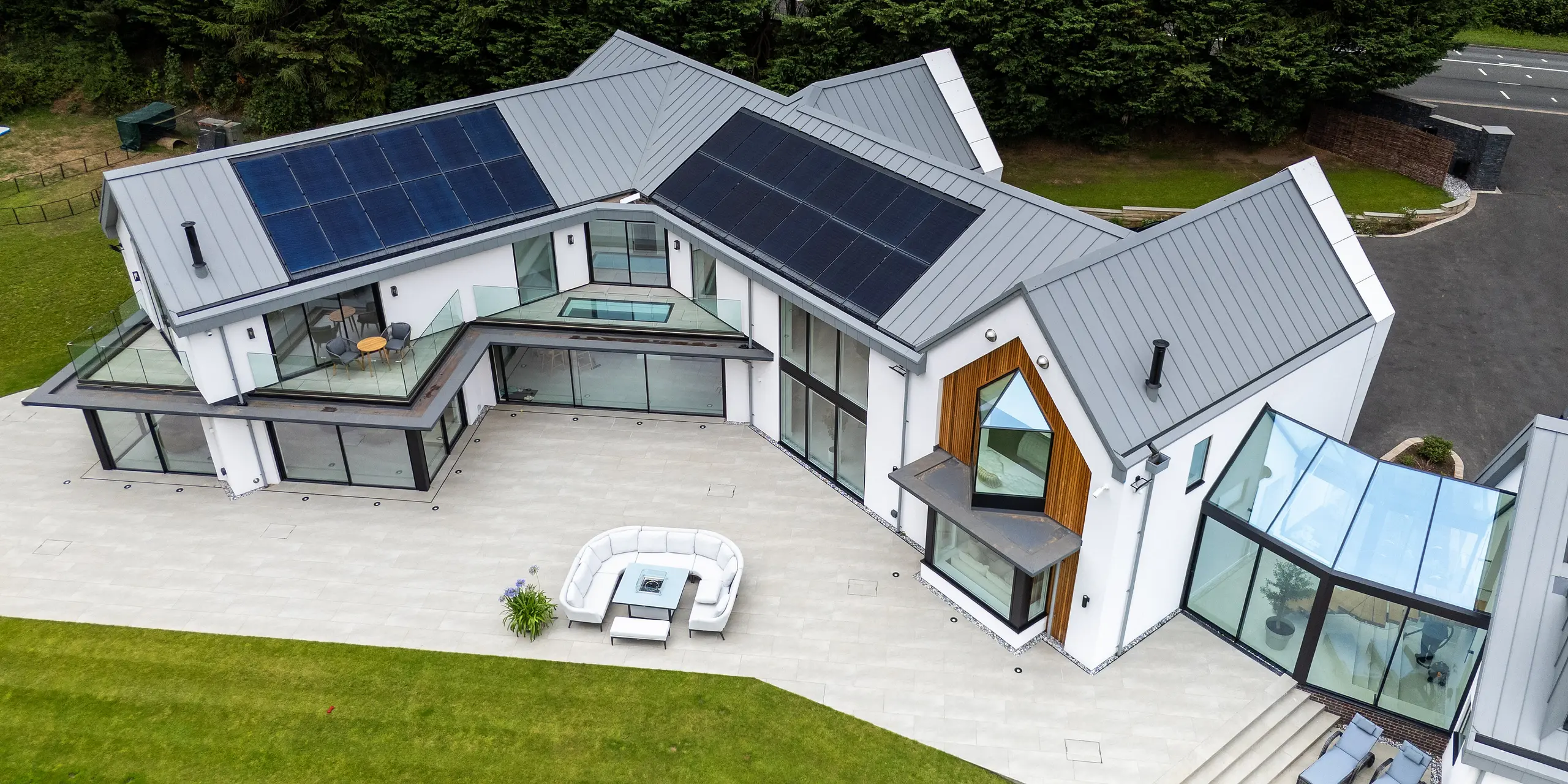 Die Ansicht von oben zeigt die beeindruckende moderne Villa in Draycott In The Clay mit ihrem PREFALZ-Dach in Patinagrau. Das Dach, mit klaren Stehfalzstrukturen und integrierter PV-Anlage, bietet sowohl ästhetische Raffinesse als auch hohe Energieeffizienz. Die große Glasfassade und die markante Holzverkleidung an der Ecke des Hauses heben die architektonische Komplexität hervor. Die weitläufige Terrasse und die gepflegte Gartenlandschaft verleihen dem Gebäude eine harmonische Verbindung zur Natur, während das PREFALZ Aluminiumdach höchsten Schutz und Langlebigkeit garantiert.