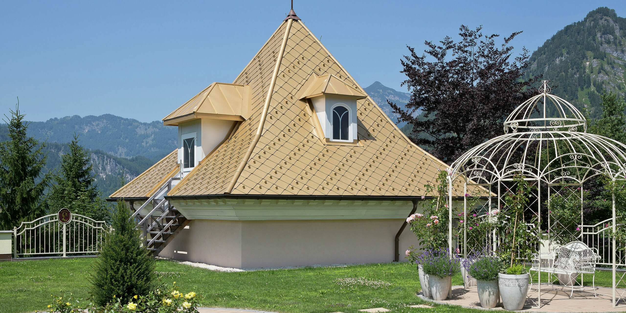 Kleiner Zubau eines Einfamilienhauses in Tirol mit PREFA Dachrauten in Mayagold und einem malerischen Garten im Vordergrund, umgeben von den Alpen.