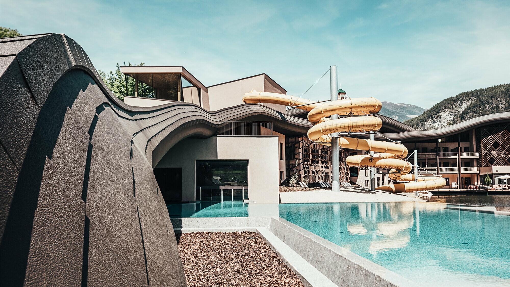  View of the pool at the Falkensteiner Hotel, a yellow water slide can be seen in the background. The roof wave of PREFA facade shingles in P.10 brown runs across the entire complex.