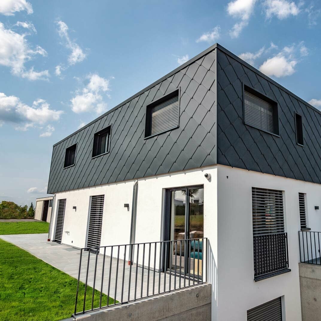 Modern new building with flat roof – the façade of the upper storey is clad with PREFA in anthracite 44 × 44 aluminium rhomboid wall tiles. The lower part of the façade is rendered in white.