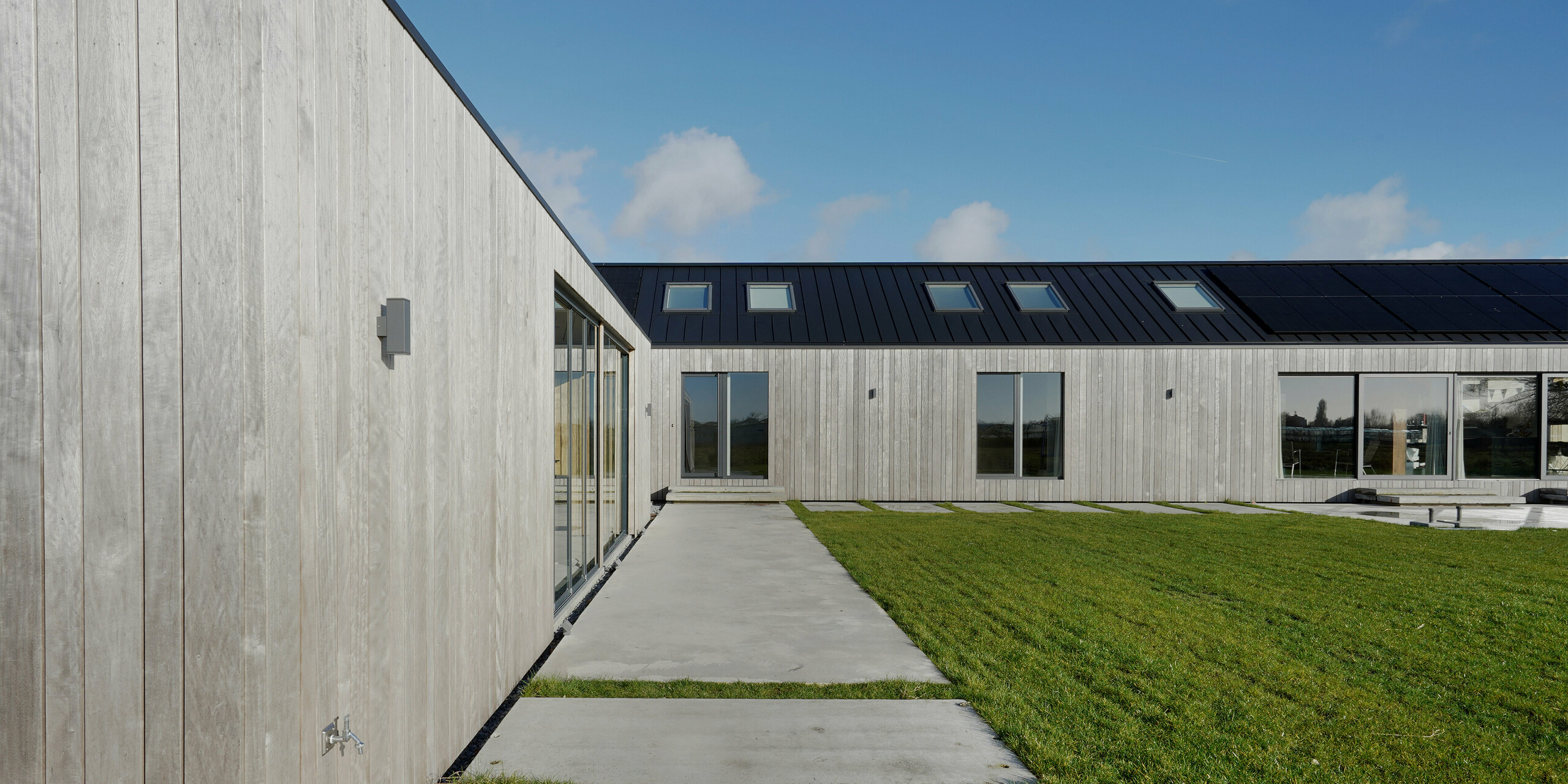 Seitliche Ansicht eines modernen Einfamilienhauses in Uithoorn, Niederlande. Das Bild hebt die natürliche Holzfassade und das präzise verarbeitete Winkelstehfalz-Dach aus PREFALZ in Schwarzgrau hervor. Der gepflasterte Gehweg entlang der Holzfassade und die großen Glasfronten des Hauses stehen zusätzlich im Fokus der Aufnahme. Die klare Linienführung und die Verwendung von langlebigem und wetterbeständigem Aluminium und nachhaltigem Holz betonen das ökologische und moderne Design des Gebäudes. Ideal für umweltbewusste Bauprojekte, die auf Qualität und Ästhetik setzen.