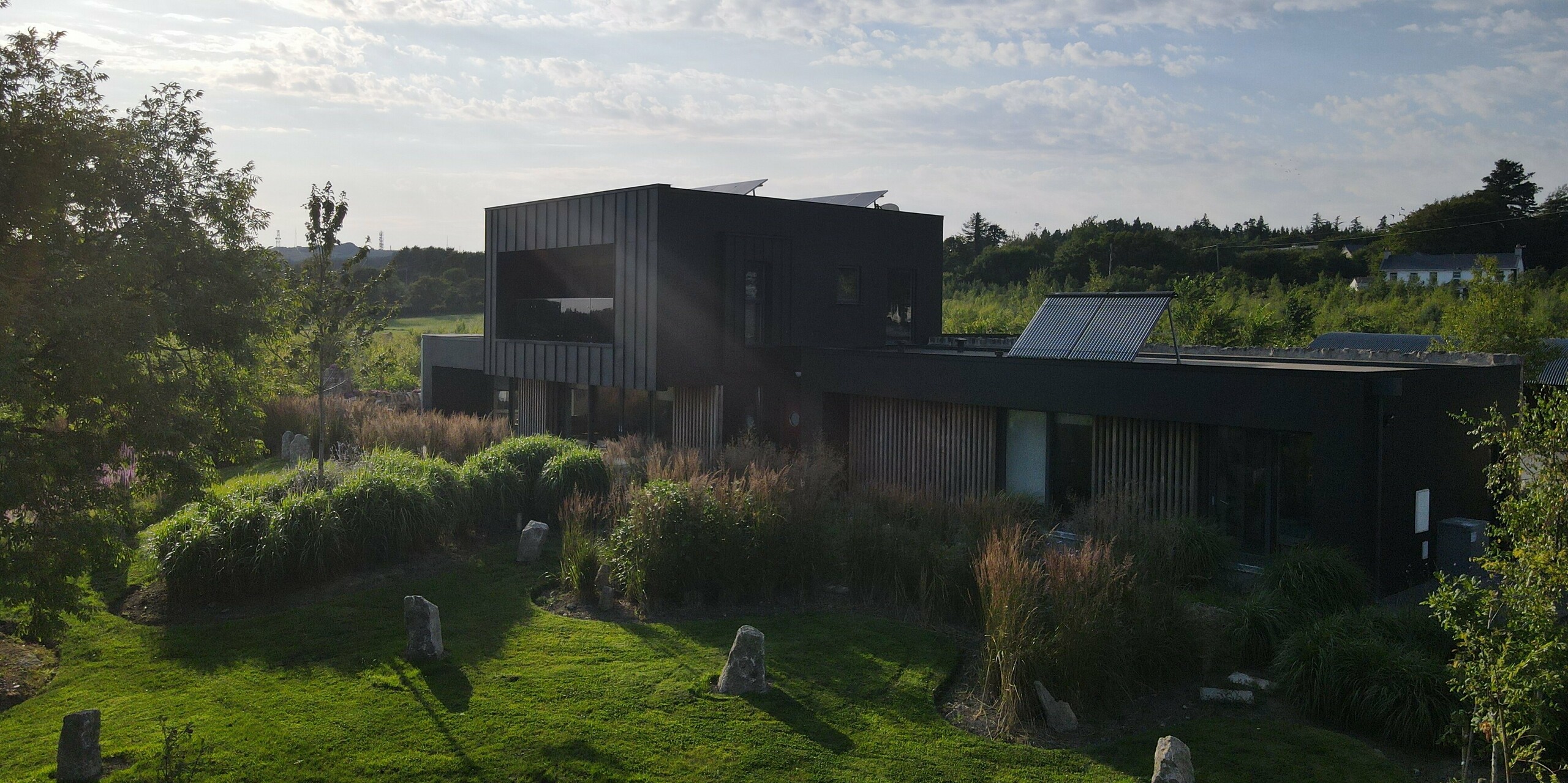 Die Aufnahme zeigt das moderne Einfamilienhaus in Wexford inmitten einer üppigen, wild gestalteten Gartenlandschaft. Besonders ins Auge fällt die schwarze PREFALZ Fassade in P.10 Schwarz, die mit ihrer klaren Linienführung und eleganten Metalloptik einen markanten Kontrast zum satten Grün bildet. Die kraftvolle Abendsonne betont die strukturierten Flächen der Aluminiumverkleidung und unterstreicht ihre hochwertige, witterungsbeständige Qualität. Das architektonische Zusammenspiel aus moderner Kubatur, großen Fensterflächen und robustem PREFA Material macht das Gebäude zu einem eindrucksvollen Highlight.