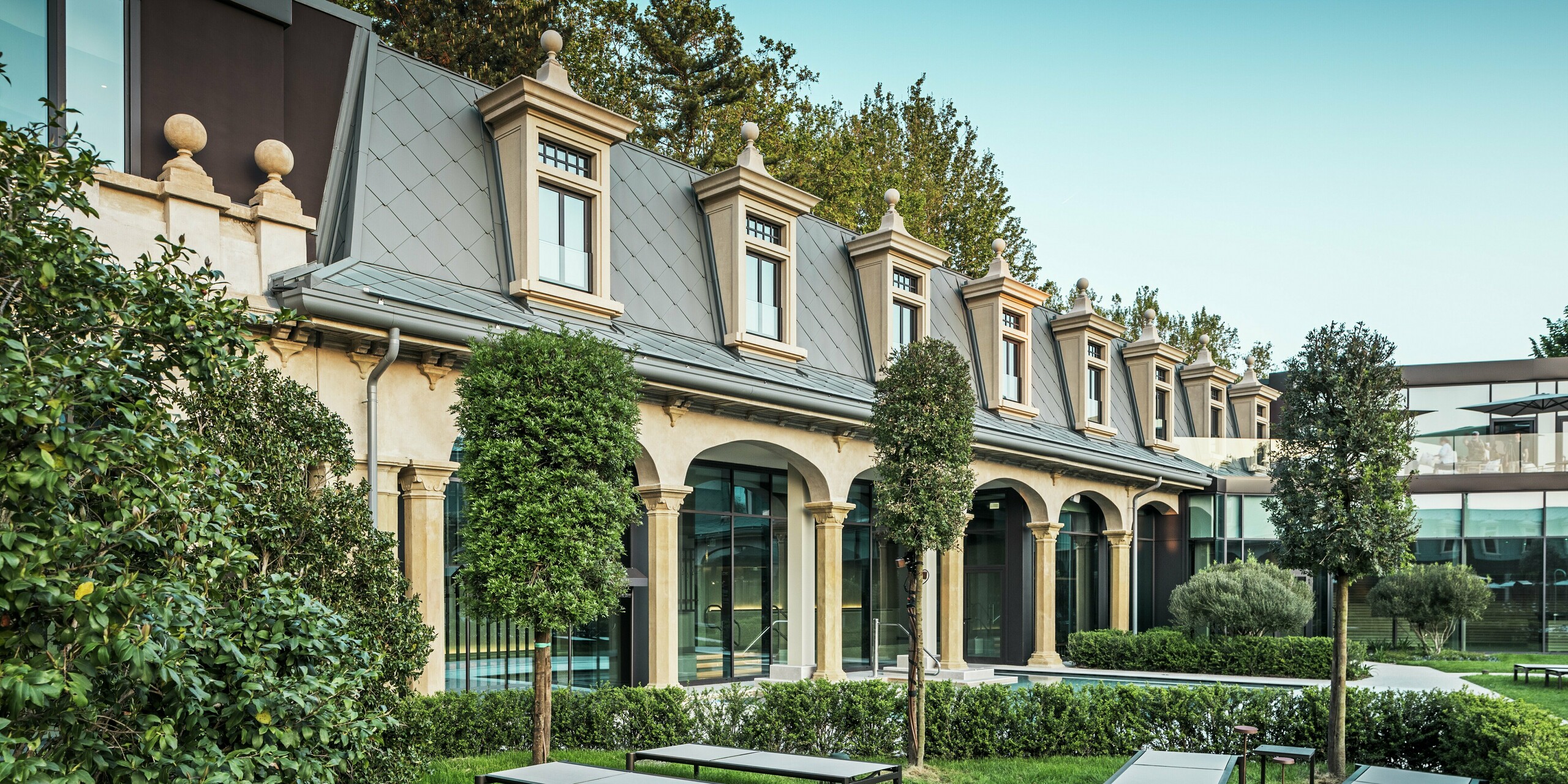 Detailansicht der stilvoll sanierten Dachlandschaft der Therme „De Montel – Terme Milano“ mit PREFA Dachrauten 44 × 44 in P.10 Hellgrau. Die präzise ausgeführten Gauben, Fensterabschlüsse und Gesimse unterstreichen die historische Eleganz des Jugendstilensembles. Im Vordergrund eine ruhige Gartenfläche mit modernen Liegen, geometrisch geschnittenen Hecken und formschönem Baumbestand – ein Ort der Erholung mit architektonischer Tiefe.