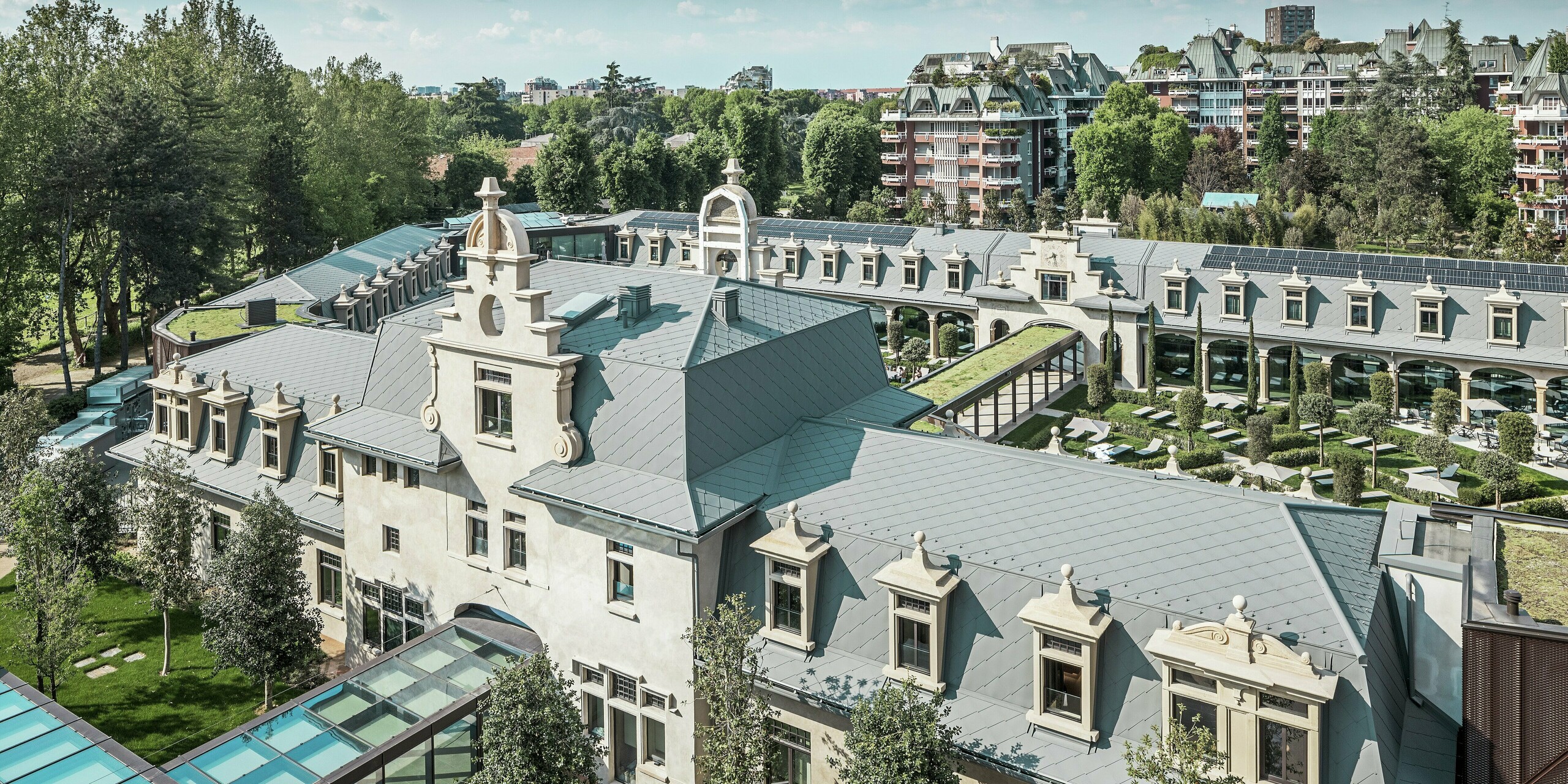 Overall view of the impressive ‘De Montel – Terme Milano’ complex with its listed Art Nouveau architecture and modern wellness ambience. The large-scale roof covering with PREFA rhomboid roof tile 44 × 44 in P.10 light grey gives the complex an elegant uniformity. Integrated PV modules, green roofs and glass connections between the buildings reflect the demand for sustainability and contemporary design in an urban context.