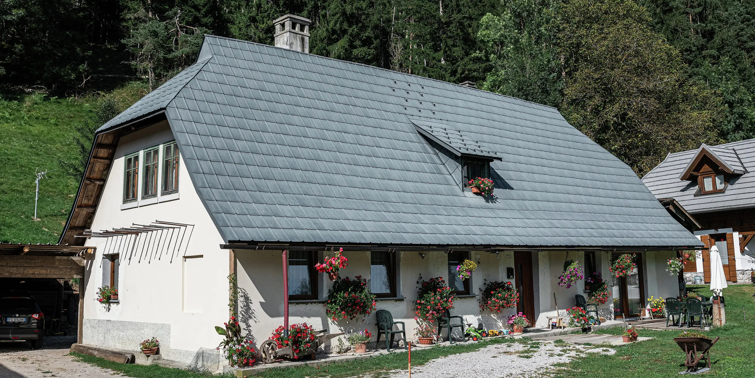 Ein idyllisches Einfamilienhaus in Tarvis, Italien, ist seit 1972 mit langlebigen PREFA Dachplatten eingedeckt. Das großflächige, witterungsbeständige Krüppelwalmdach aus Aluminium schützt das Haus seit Jahrzehnten vor den alpinen Witterungsbedingungen. Die Fassade ist mit zahlreichen Blumen geschmückt, die für eine einladende Atmosphäre sorgen. Eine kleine Terrasse mit Sitzgelegenheiten lädt zum Verweilen ein. Im Hintergrund erstreckt sich ein dichter Wald, der das Haus harmonisch in die Natur einbettet. Links ist eine überdachte Einfahrt mit einem geparkten Auto zu sehen, während rechts ein weiteres charmantes Holzhaus das Bild abrundet.