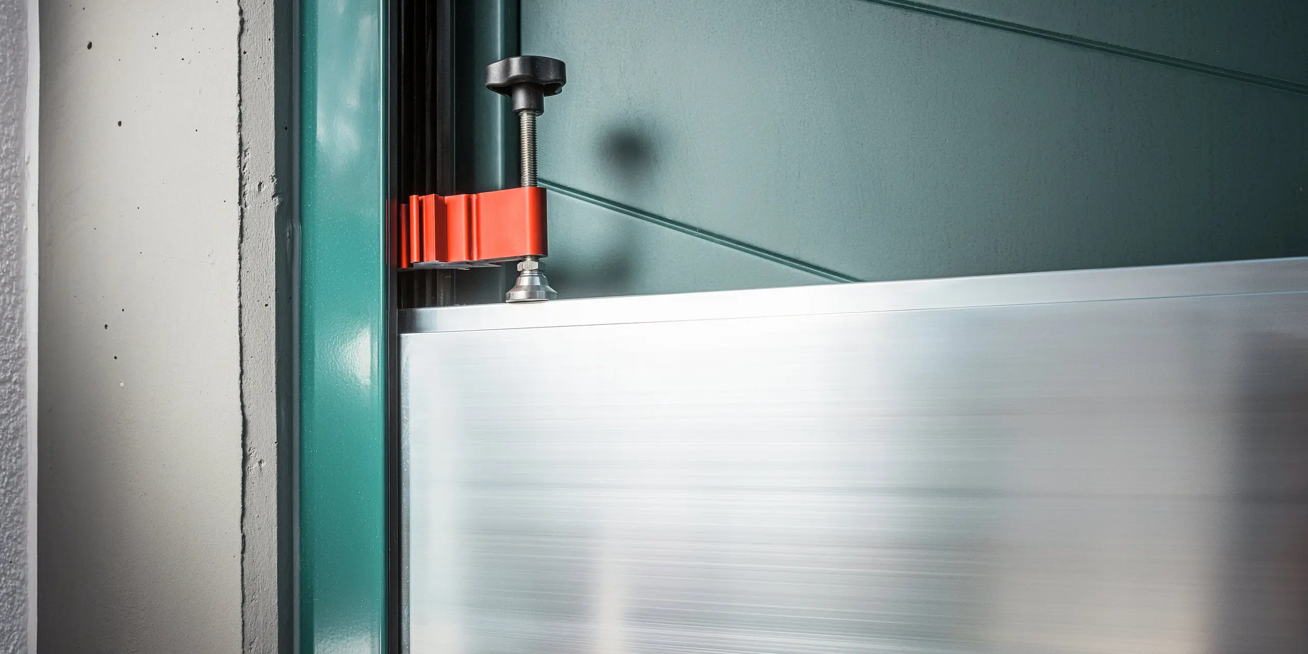 Nahaufnahme der Spannvorrichtung des PREFA Hochwasserschutzsystems im Türbereich eines Einfamilienhauses in Schwaz. Zu sehen ist die präzise Fixierung der Aluminium Dammbalken in den seitlichen Wandprofilen mittels rotem Spannbügel und schwarzer Schraubklemme. Die hochwertige Verarbeitung und die einfache Handhabung unterstreichen die Alltagstauglichkeit des Systems - ideal für den schnellen Hochwasserschutz an Hauseingängen.